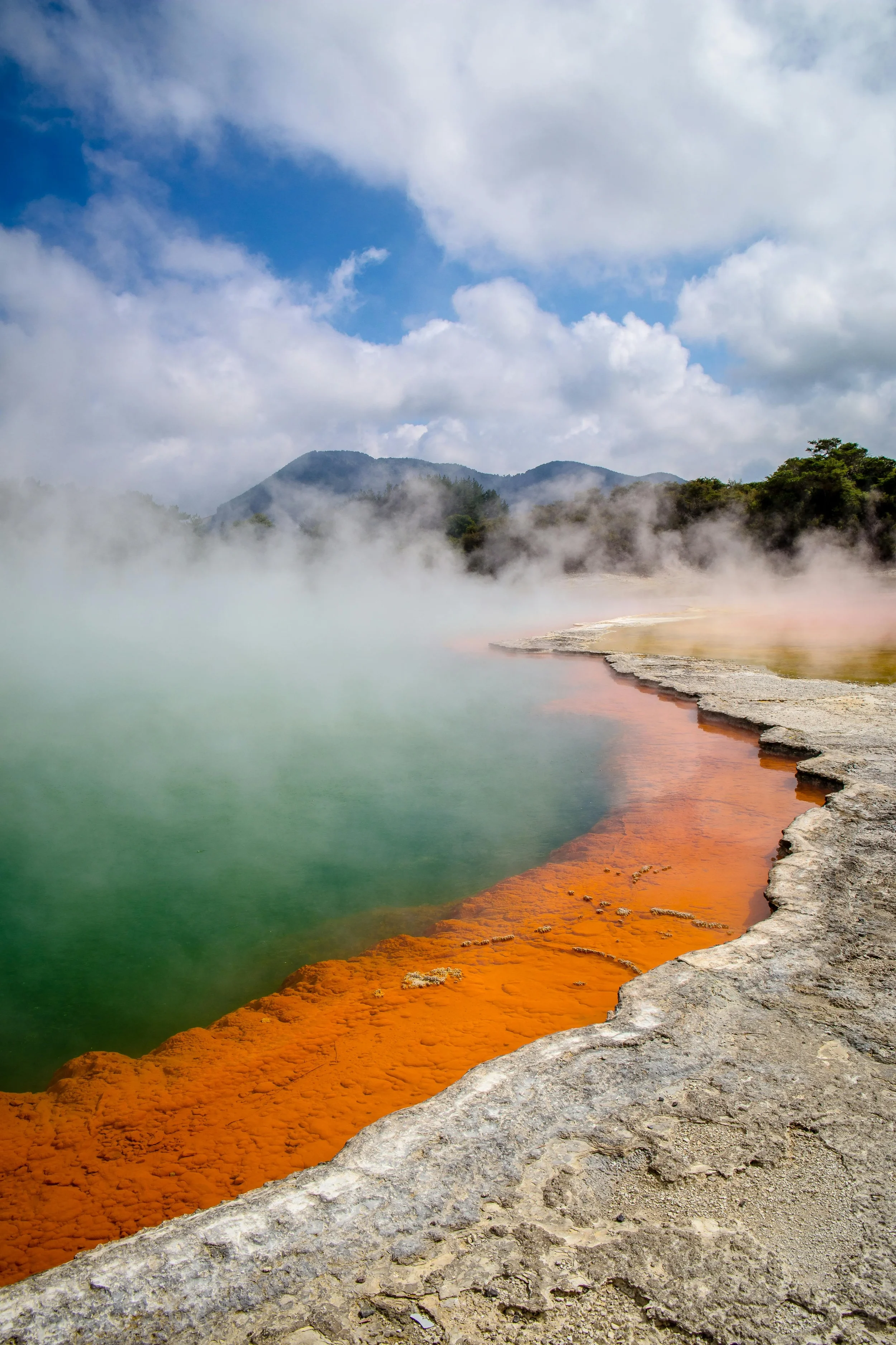 Steam rising from a geothermal hot spring with vibrant orange, green, and yellow mineral deposits along the edge, set against a backdrop of mountains and a cloudy sky.