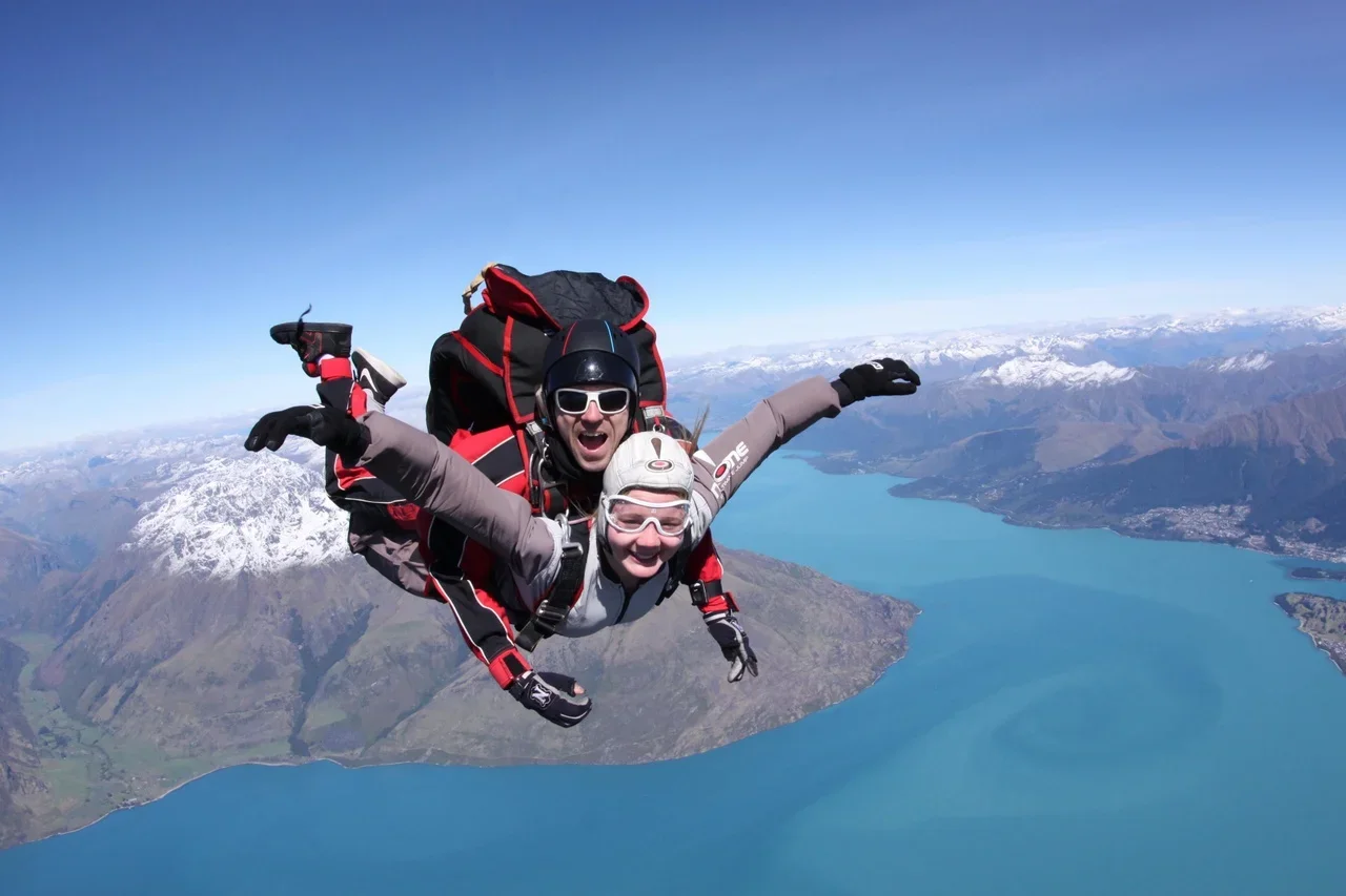 Two skydivers, wearing helmets and jumpsuits, free-falling above a body of water with mountains in the background.