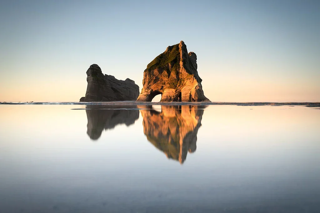 A large rock formation in the ocean with a natural arch, reflecting in the water at sunrise or sunset with a clear sky.
