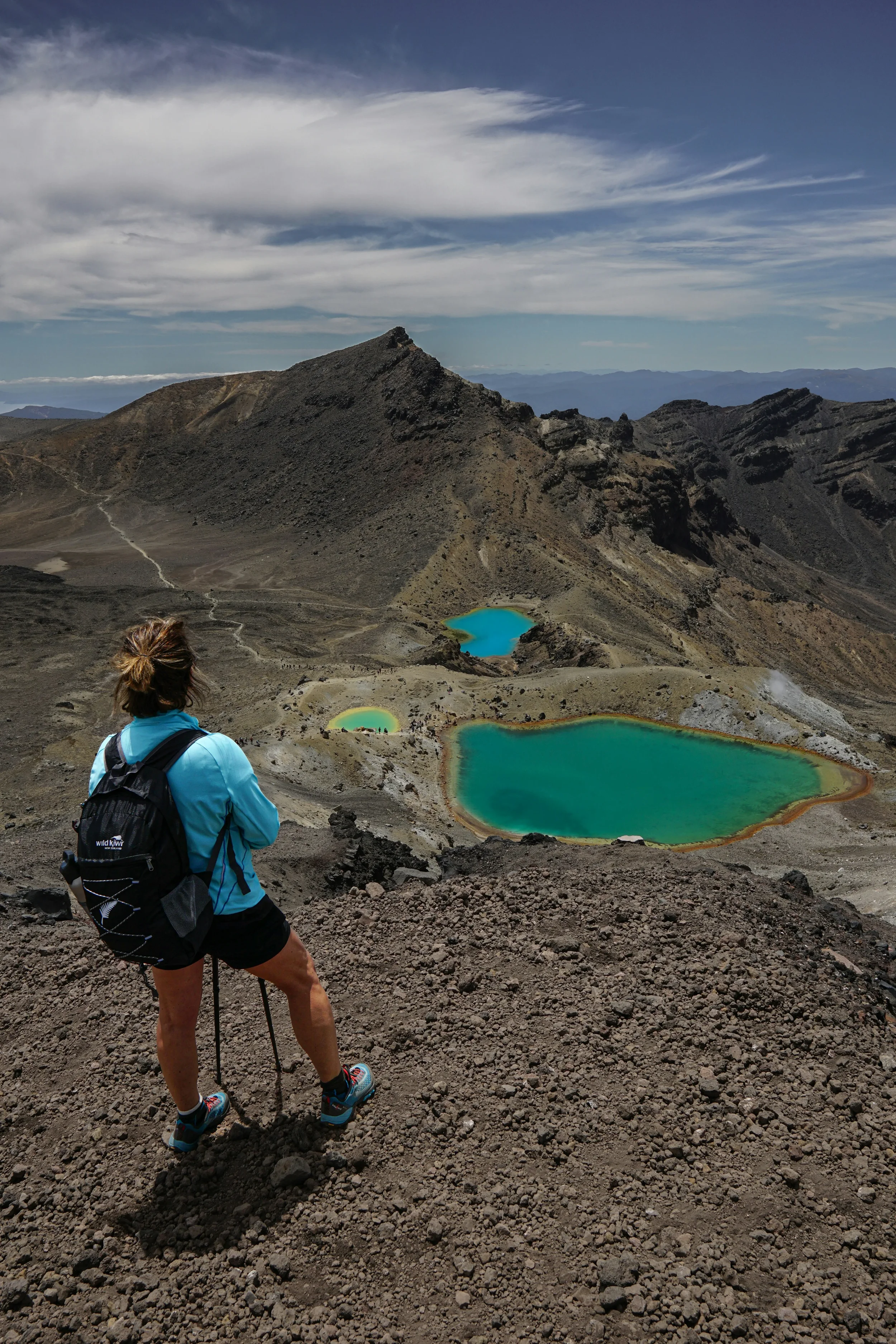 Hiker wearing a blue jacket, black shorts, and a backpack, standing on rocky terrain overlooking a volcanic crater with colorful lakes and rugged volcanic mountains in the background under a blue sky with clouds.