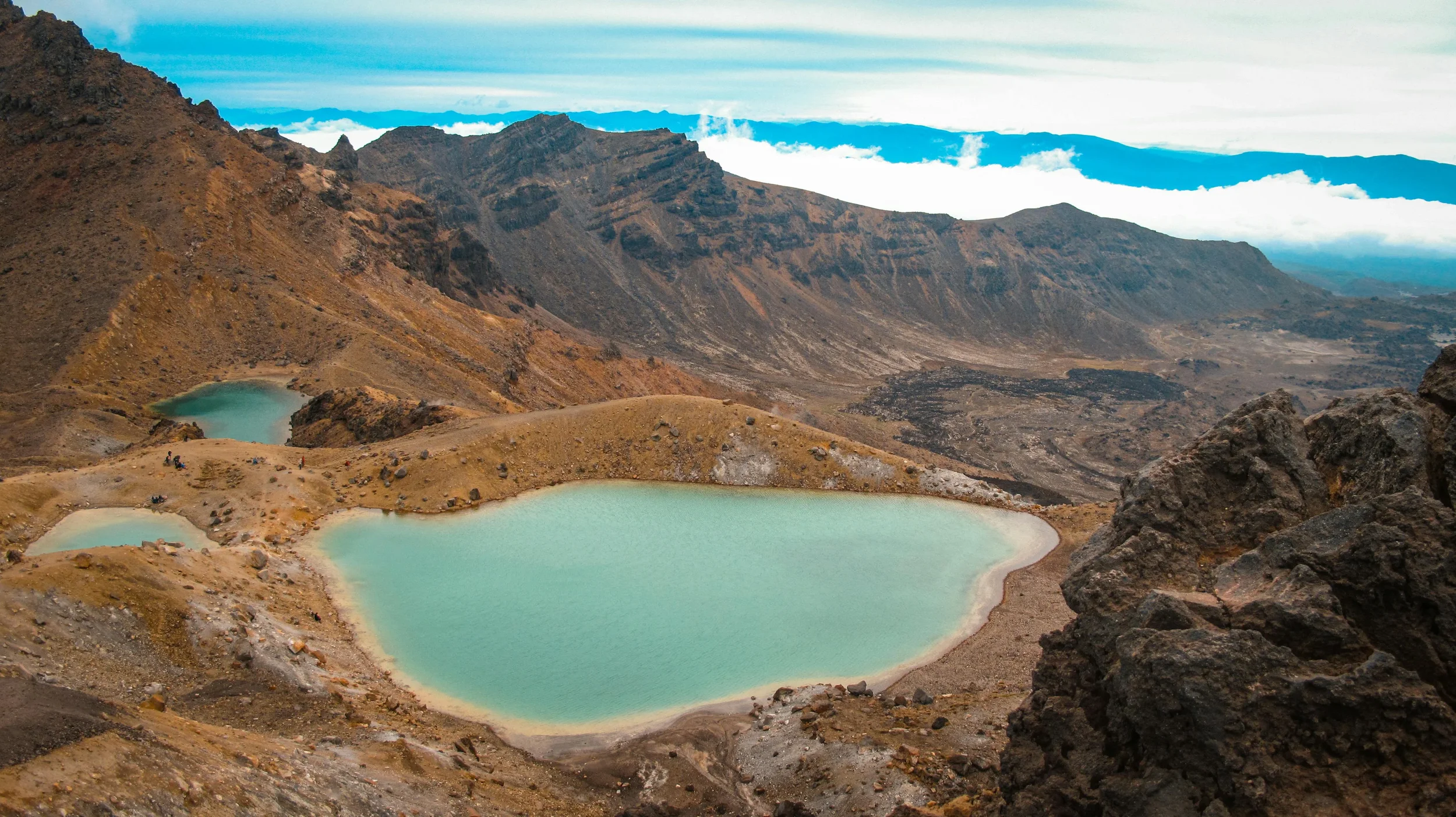 Volcanic crater with four turquoise lakes surrounded by rocky, barren terrain, mountain peaks in the background, and a cloudy sky overhead.