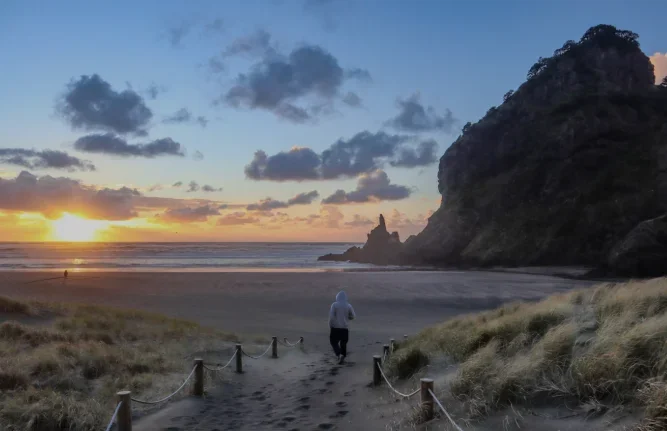 A person walking on a sandy path towards a beach during sunset, with clouds in the sky and a large rocky hill on the right.