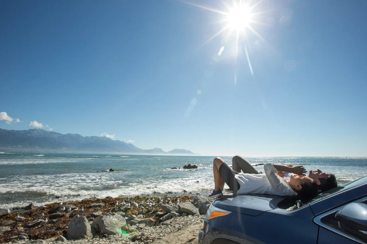 Two people lying on the hood of a car parked on a rocky beach, enjoying the sunny weather with ocean waves and mountains in the background.