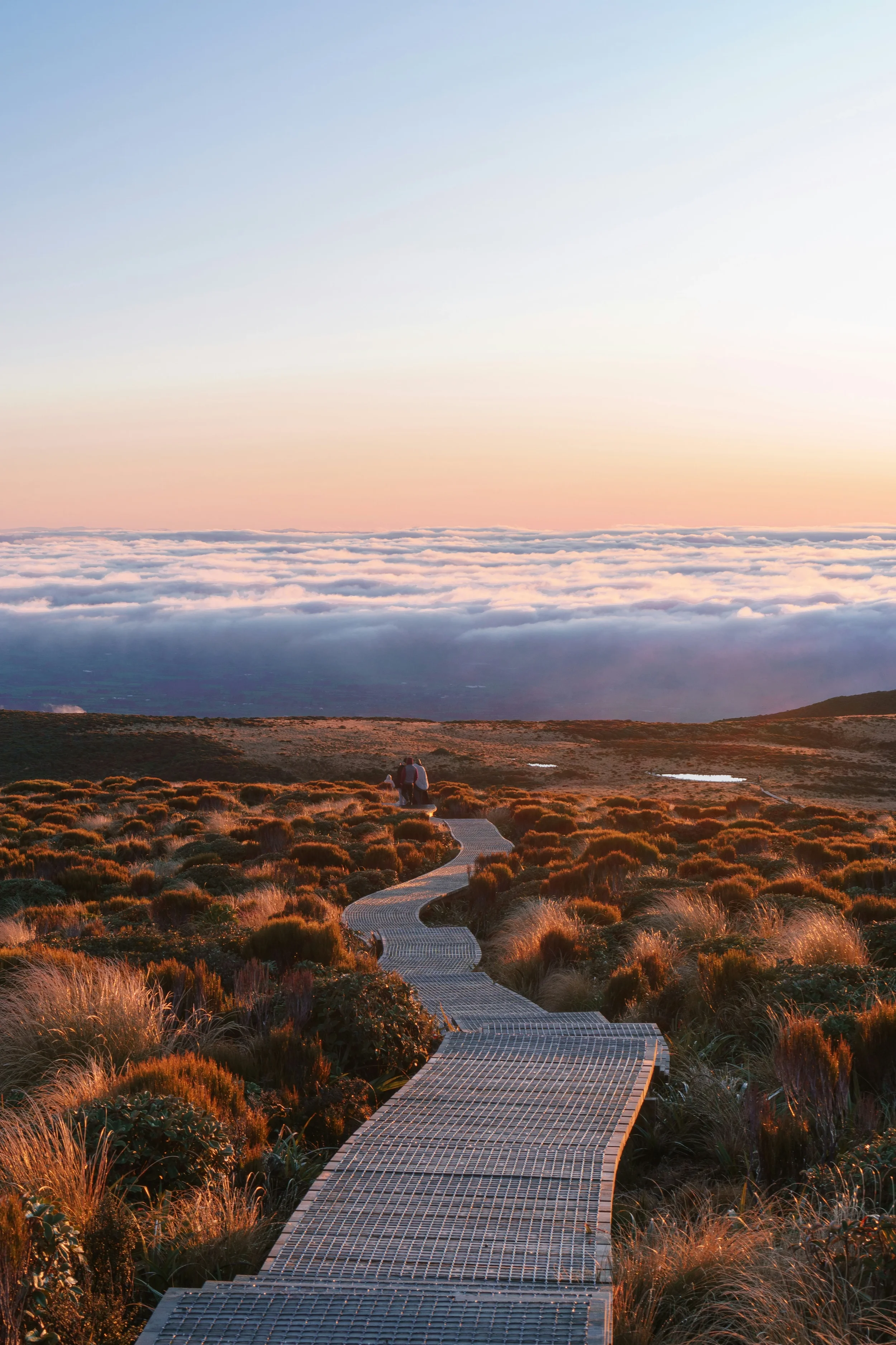 A winding path through grassy hills during sunset with a view of clouds below in the sky.