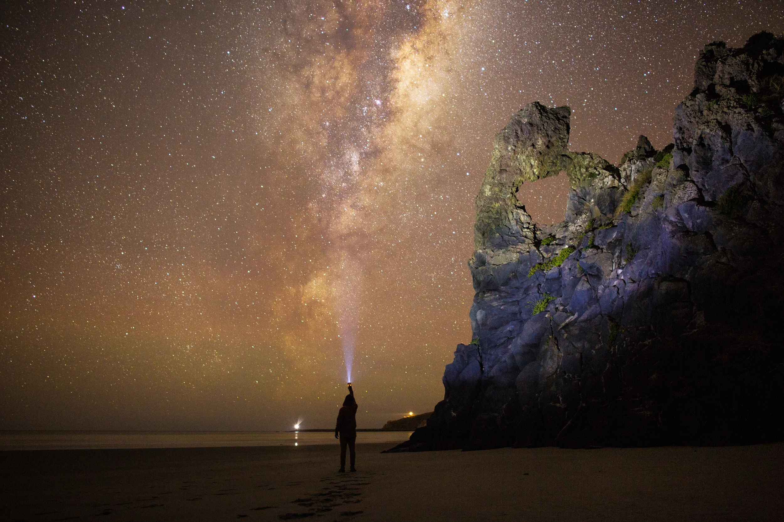 A person standing on a beach at night, looking at the starry sky and the Milky Way galaxy, with rocky cliffs and a lighthouse shining in the distance.