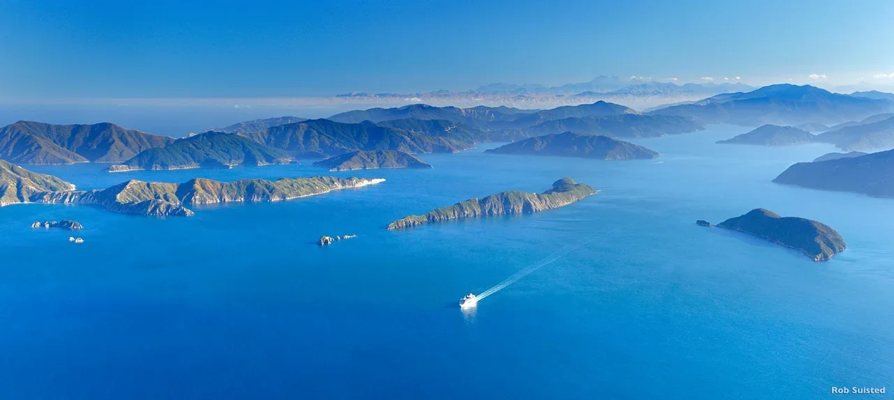 Aerial view of a large blue body of water with several green islands and mountains in the background, with a boat leaving a trail behind.