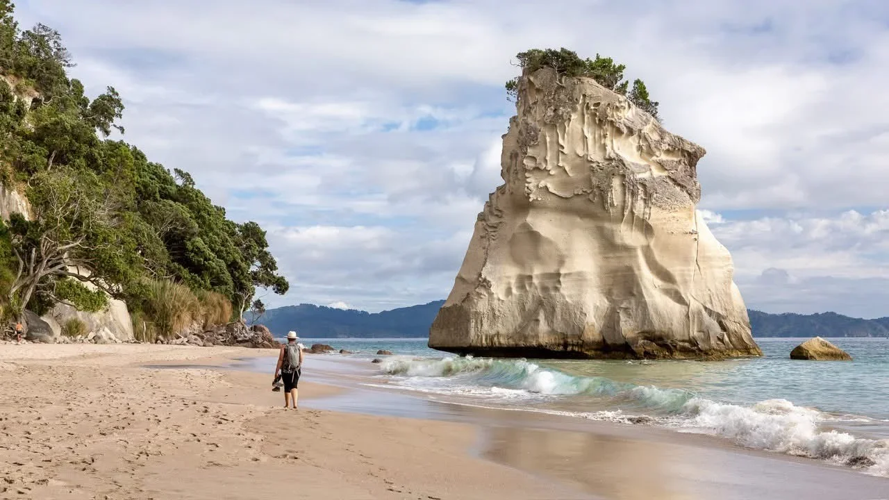A person walking on a sandy beach with lush trees on the left and a large rock formation in the water on the right, under a partly cloudy sky.