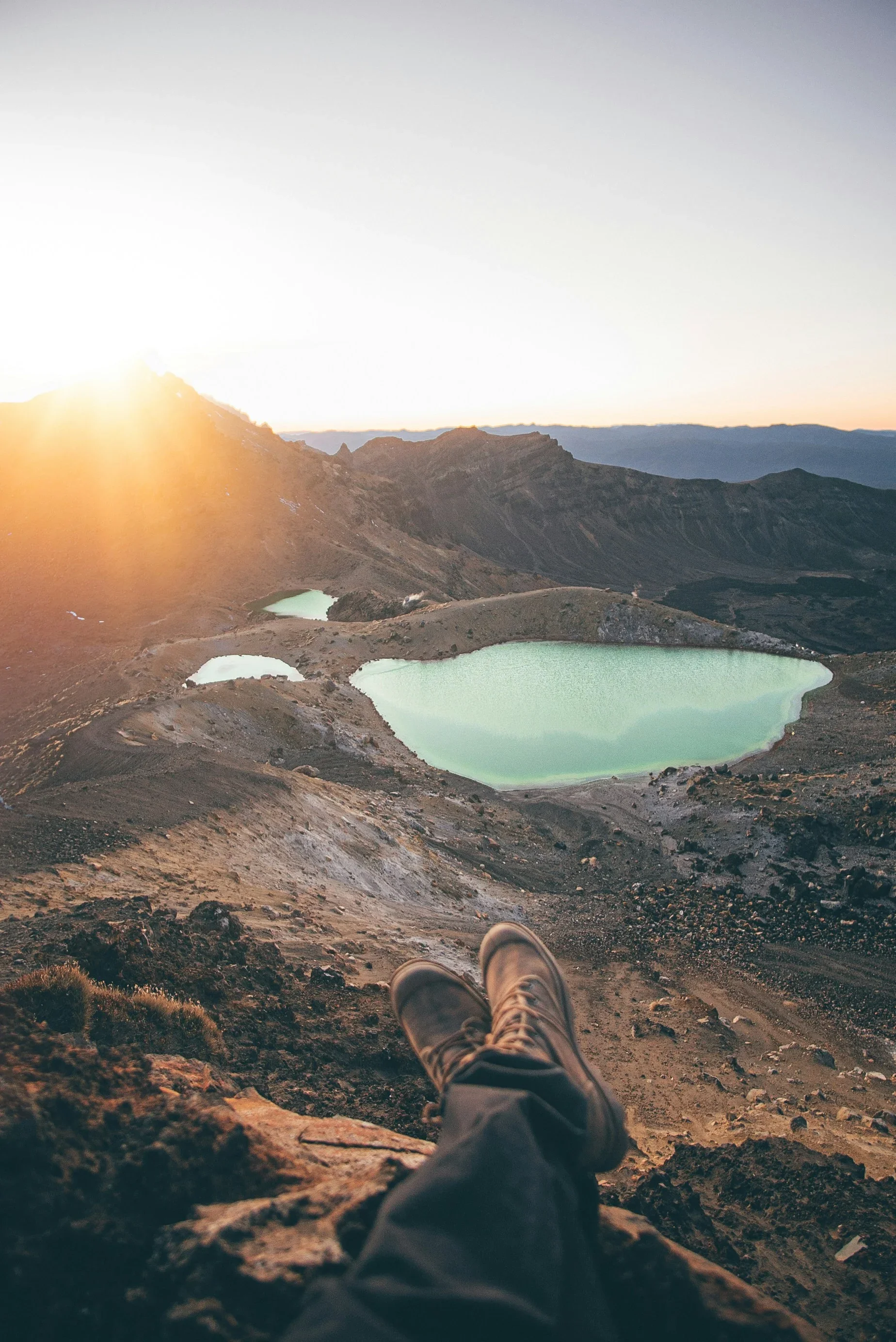 Person sitting with legs extended on rocky terrain overlooking a mountain landscape with a lake, sunrise, and a distant mountain range.