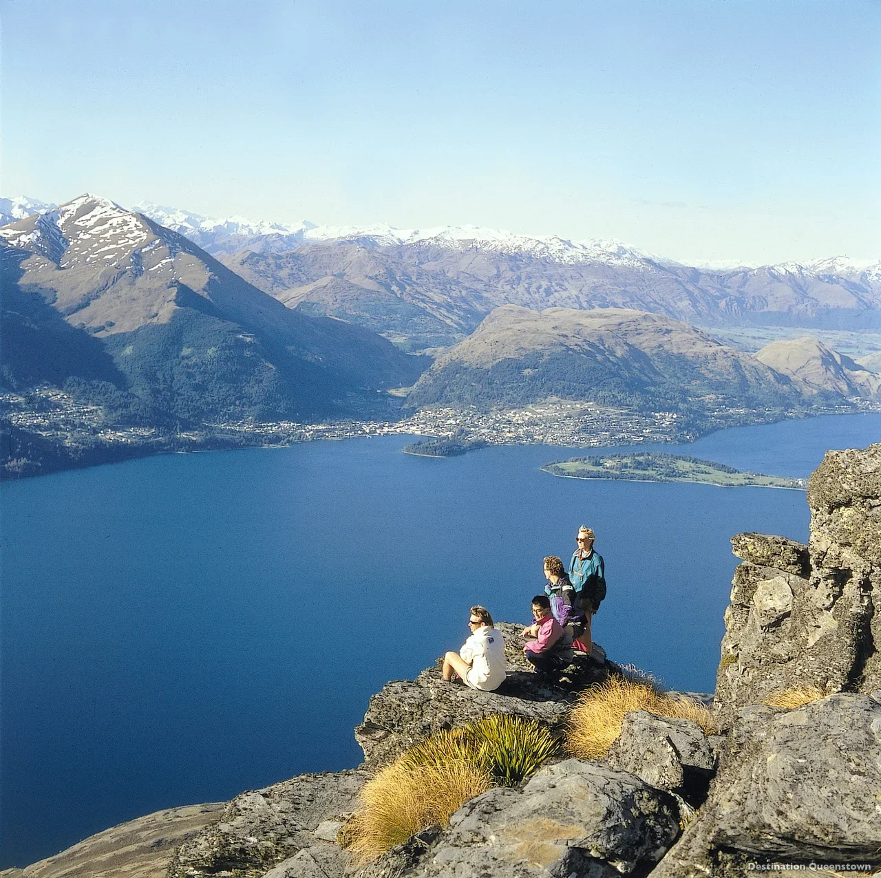 Four people sitting and standing on a rocky ledge overlooking a large lake, mountain range with snow-capped peaks in the background, and a clear blue sky.