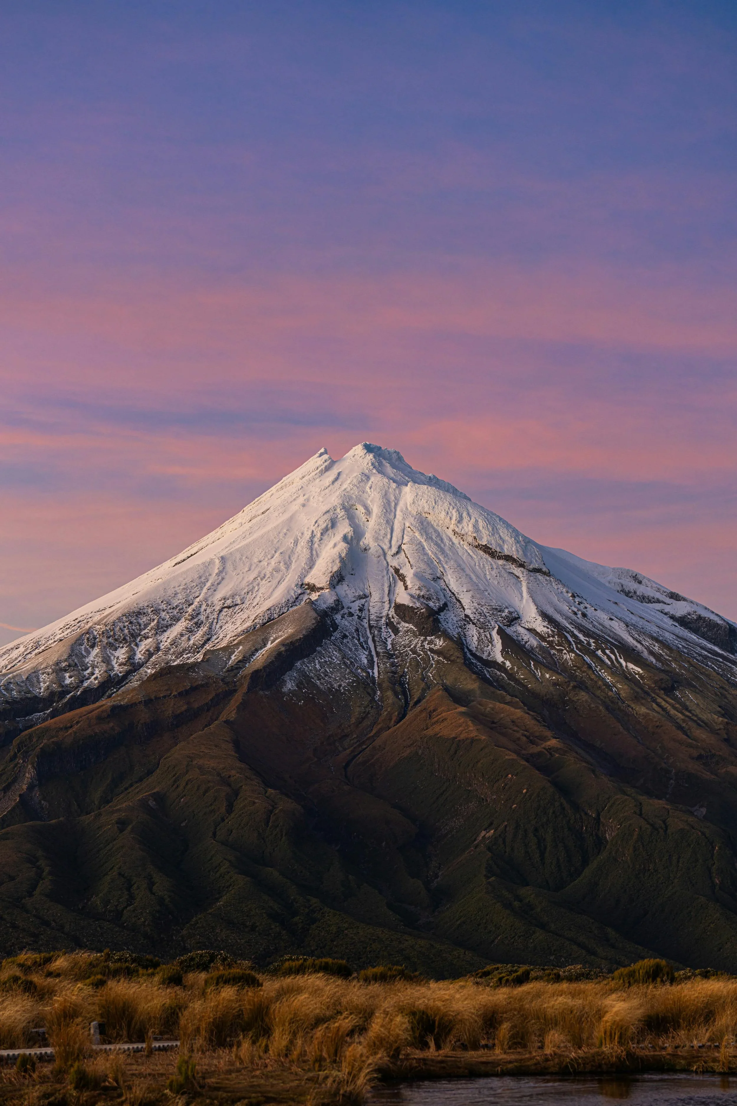 A snow-capped mountain under a colorful sky with clouds, with green hills and a body of water at the foreground.