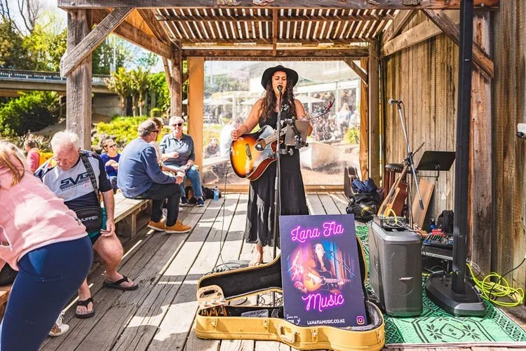 A woman singing and playing guitar at an outdoor wooden stage with a microphone, surrounded by an audience seated on benches, with music equipment and a sign reading 'Lana Fia Music' in front.