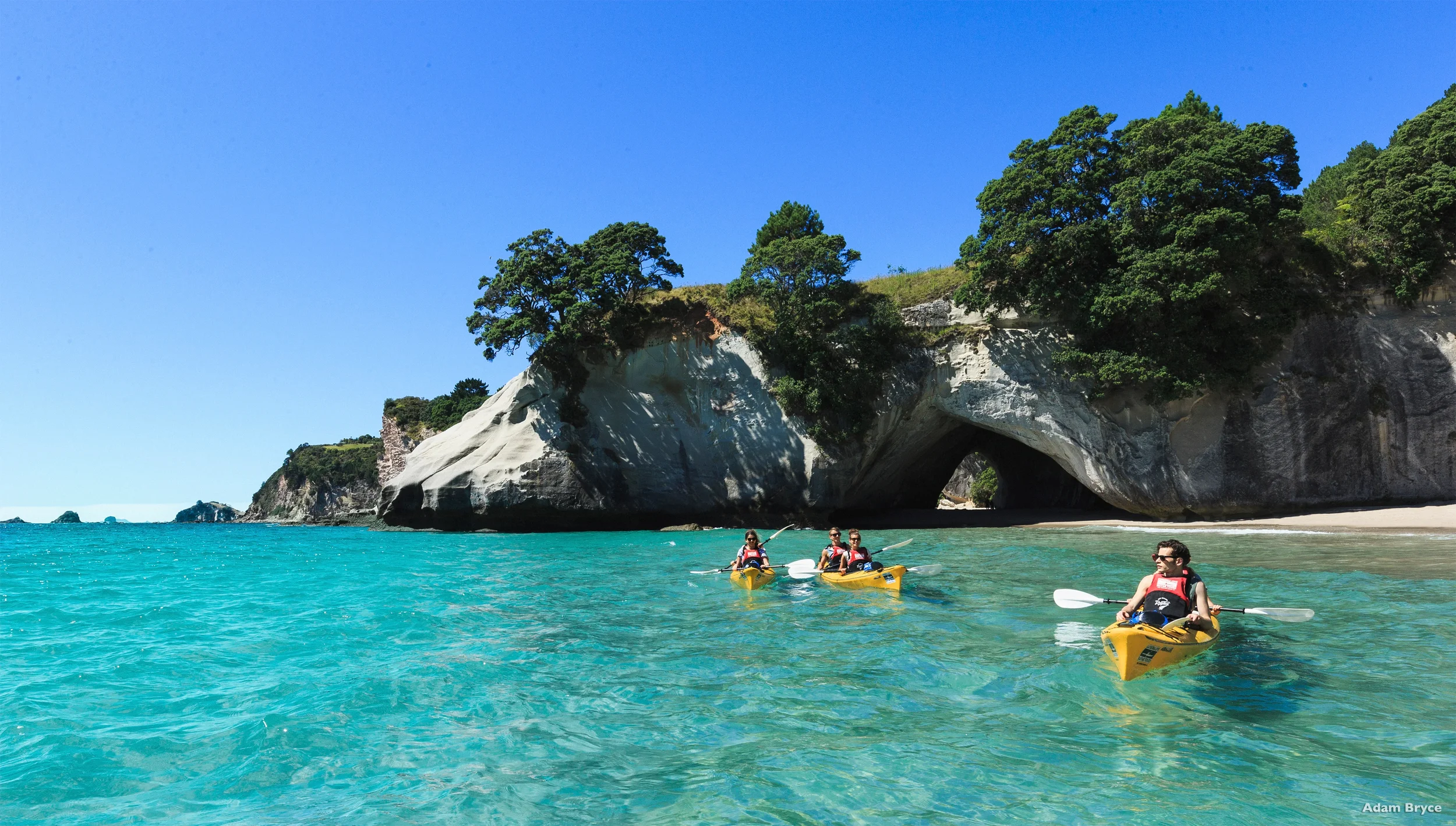 Group of people kayaking on turquoise water near a rocky cliff with lush trees and a natural arch formation under a clear blue sky.