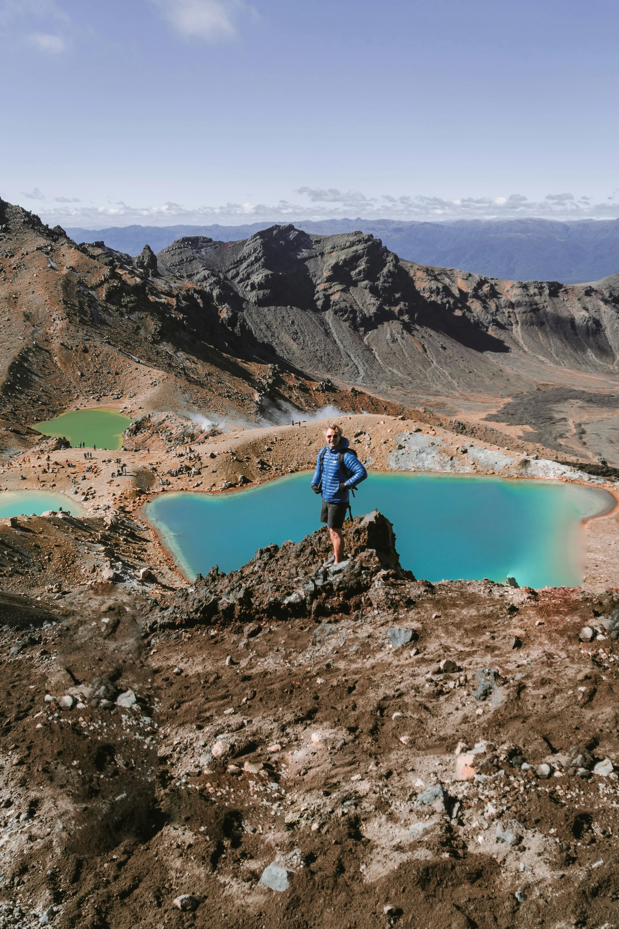 A hiker standing on rocky terrain near turquoise volcanic crater lakes, with rugged mountain landscape and blue sky in the background.