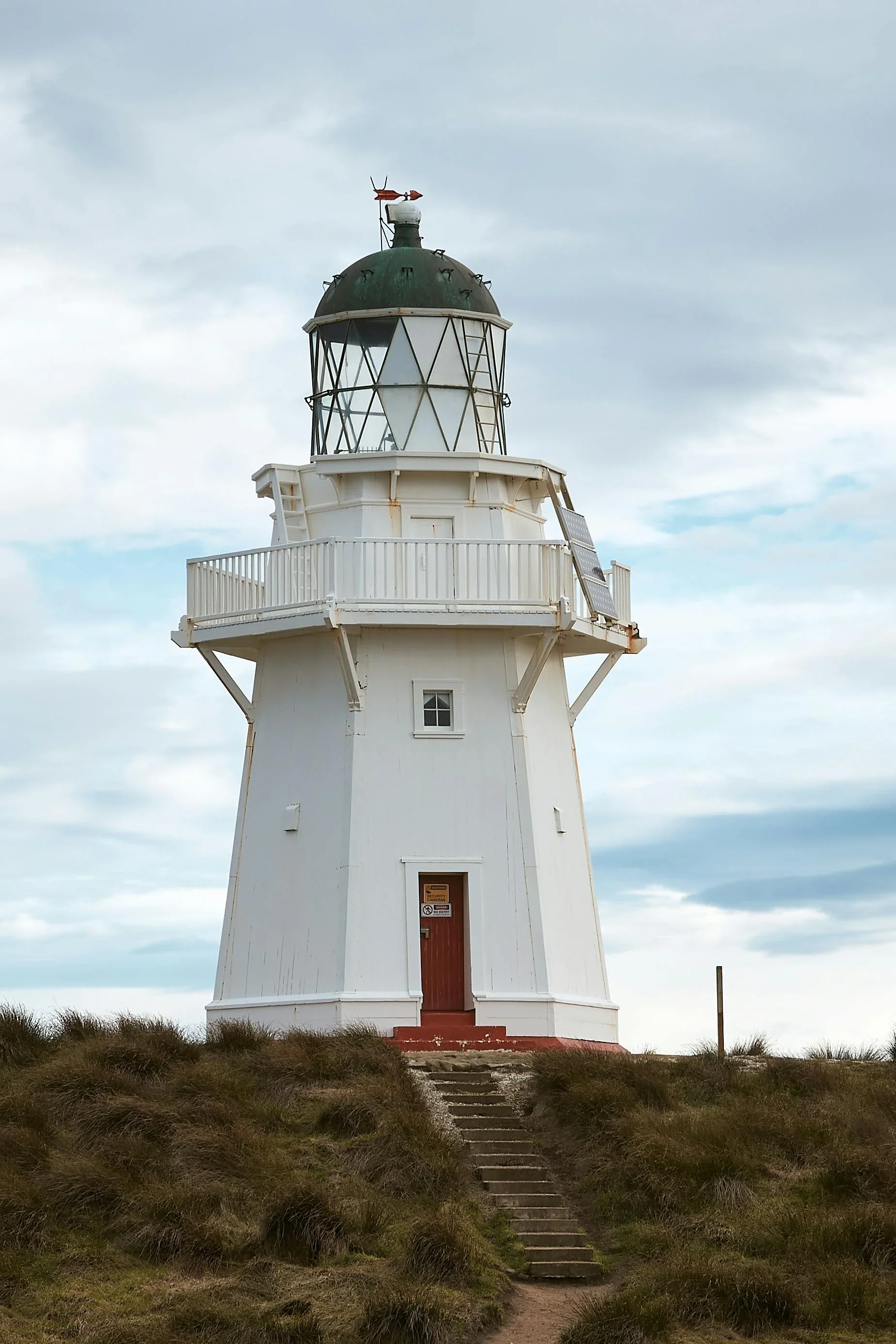 A white lighthouse with a green roof, situated on a hill with steps leading up to it, under a cloudy sky.