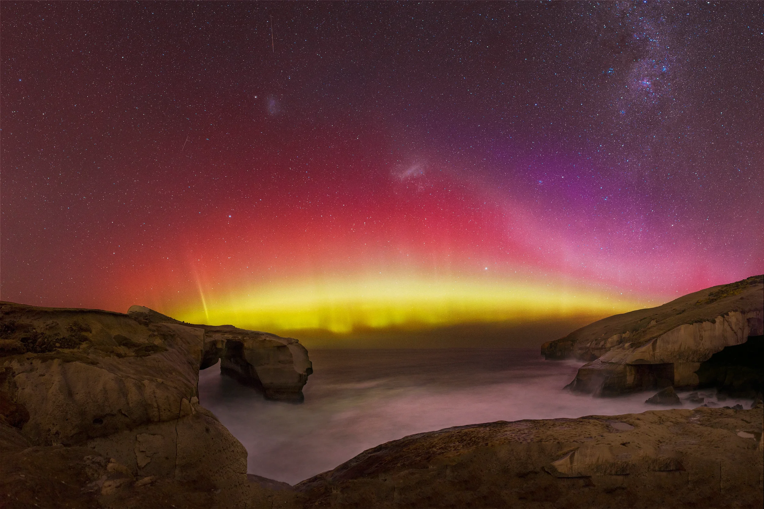 Night sky over a rugged coastline with cliffs and rock formations, displaying the Northern Lights with vibrant pink, purple, and yellow hues, and a starry sky including the Milky Way and a faint shooting star.