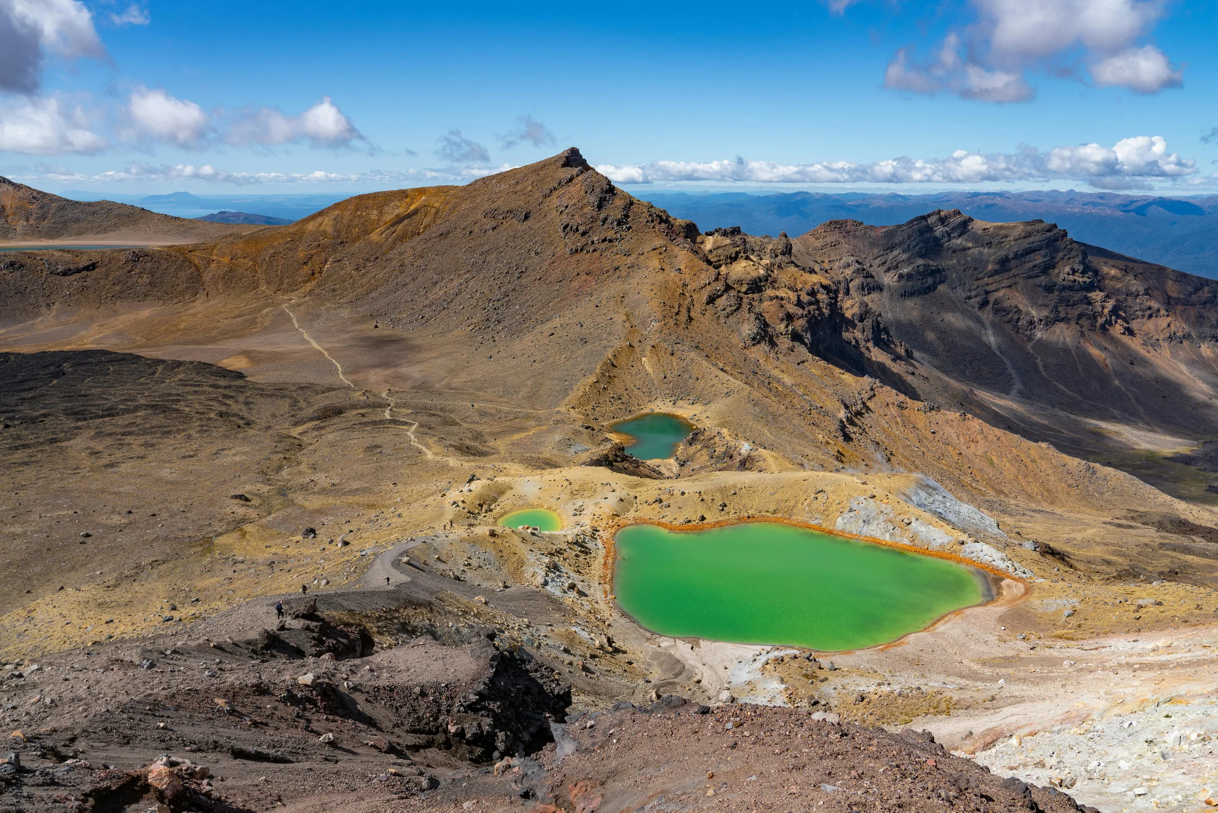 View of a volcanic landscape with rugged terrain, multiple small green crater lakes, and a mountain peak under a partly cloudy sky.