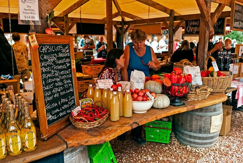 Vendor at a farmers market stall selling fresh produce like apples, pumpkins, peppers, and bottled juice. Customers browse in the background.