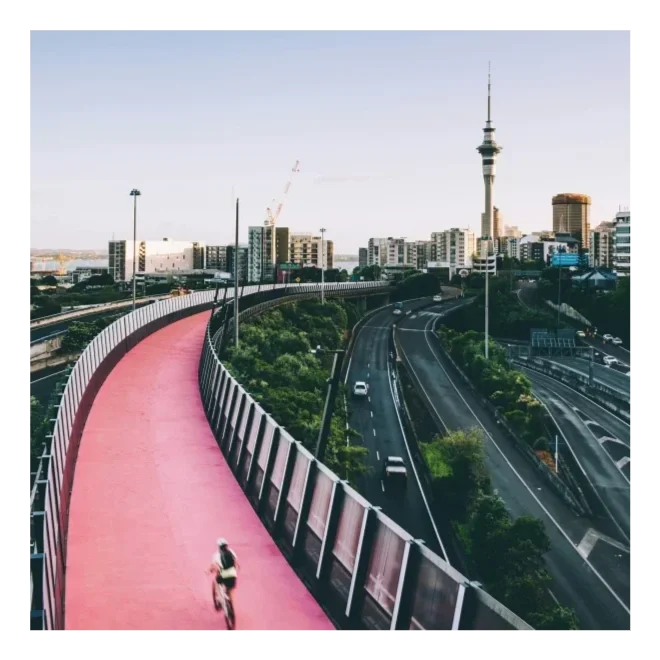 A person running on a pink elevated pathway over a city road with multiple lanes, buildings, and a tall observation tower in the background.