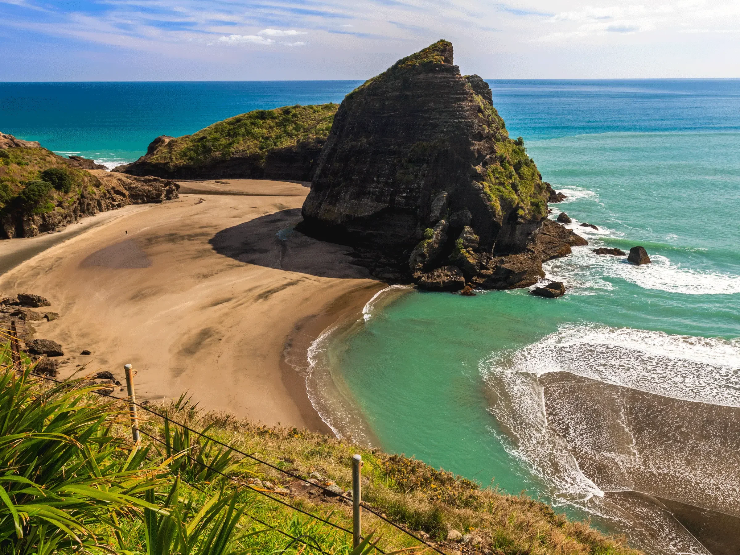 A coastal landscape with a sandy beach, a large rocky formation covered in greenery, and the ocean with blue and green waters under a partly cloudy sky.