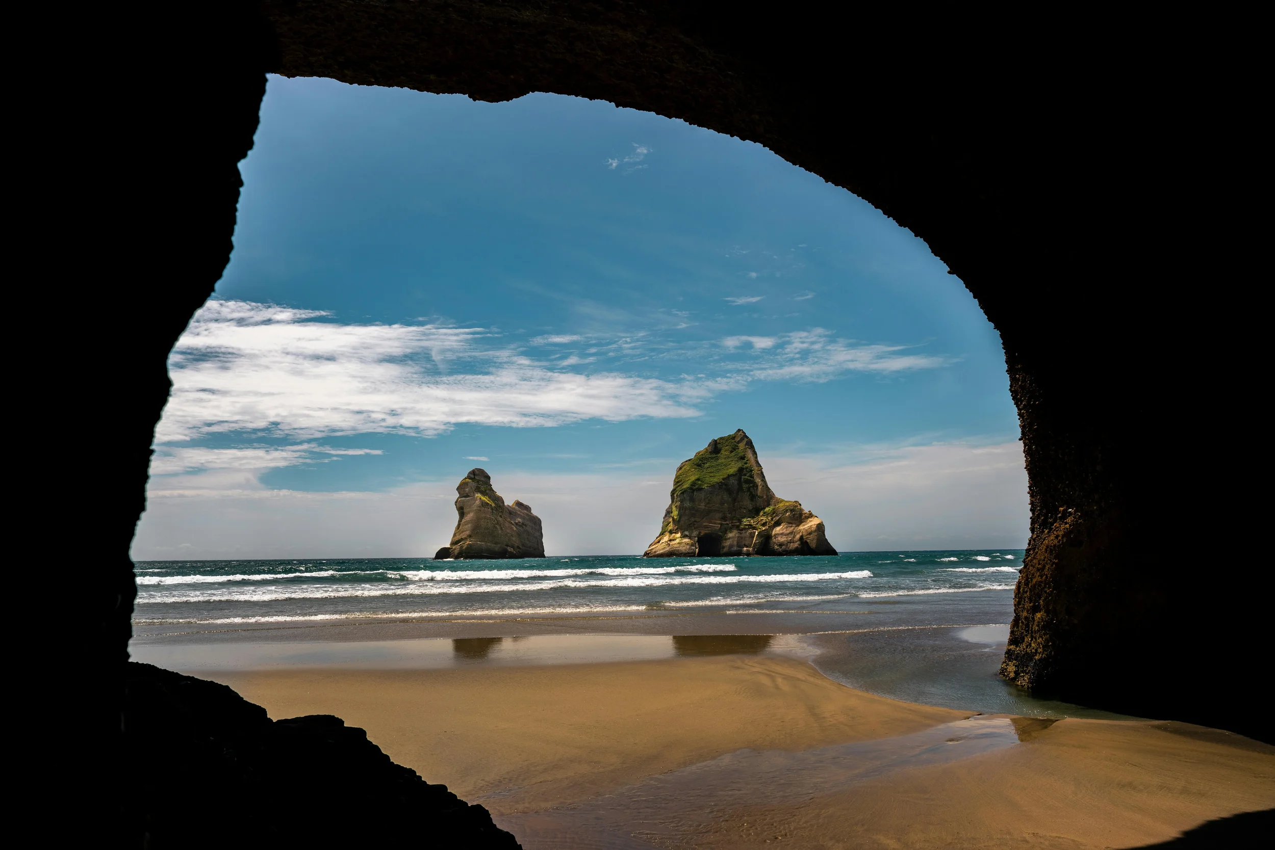 View from inside a cave looking out at a beach with large rock formations in the water, blue sky, and scattered clouds.