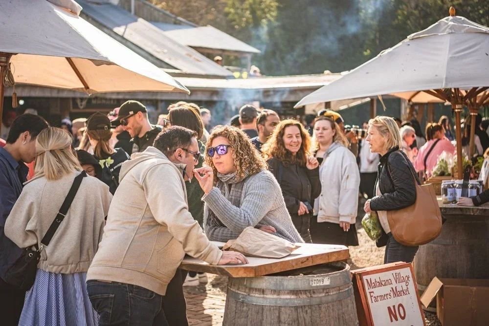 People shopping at an outdoor farmers market with umbrellas, including a woman in sunglasses and a gray sweater talking to a man, and other shoppers browsing nearby.