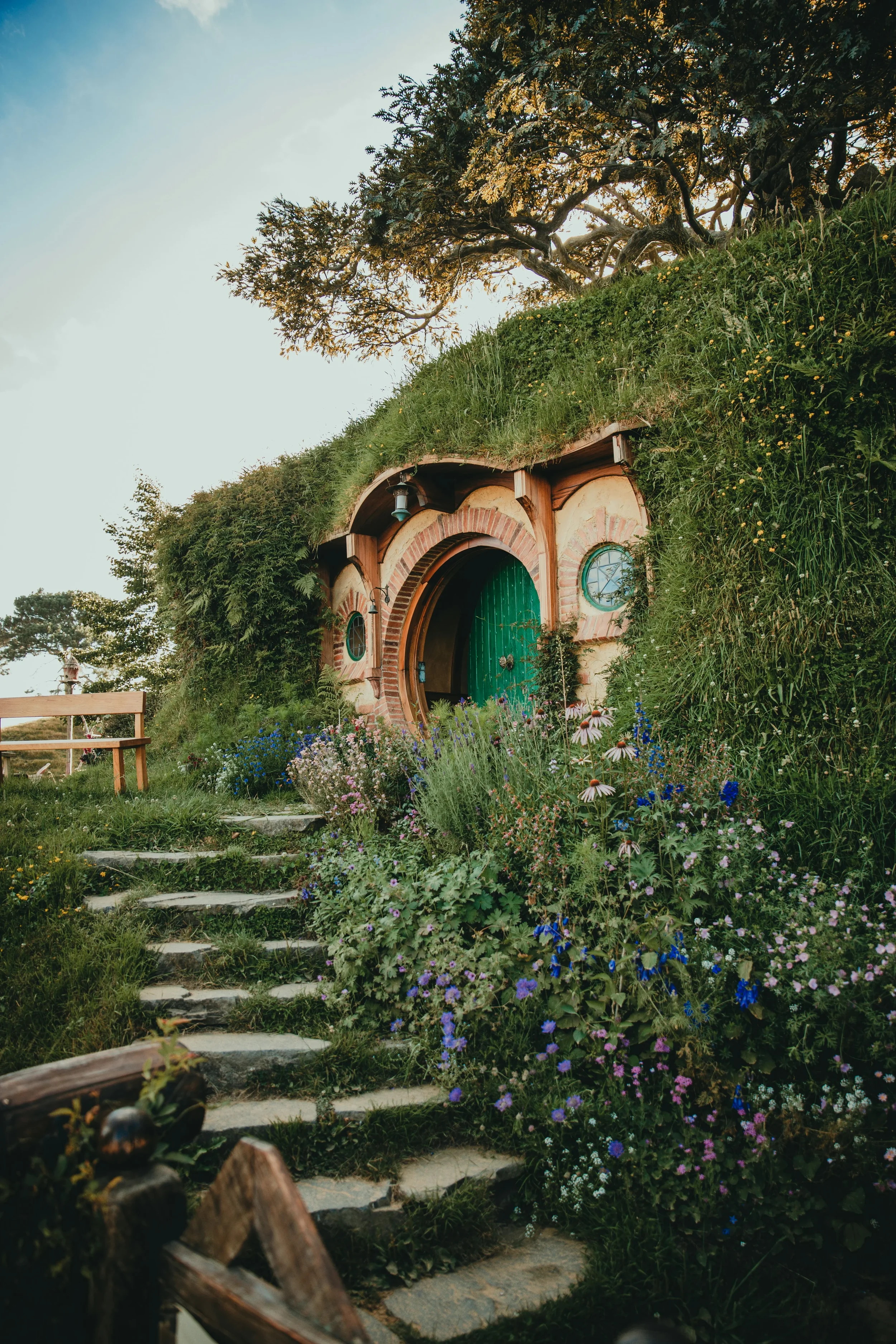 A Hobbit-style house with a circular green door, brick archway, and small windows, surrounded by colorful flowers, lush greenery, and a stone pathway, with a large tree overhead and a blue sky background.