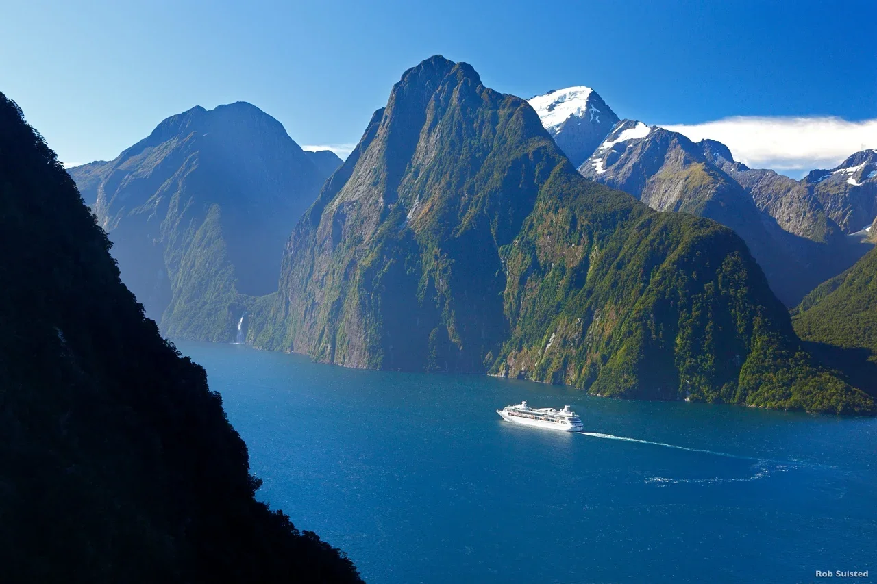 A cruise ship sailing on a deep blue fjord surrounded by towering green mountains with snowy peaks.