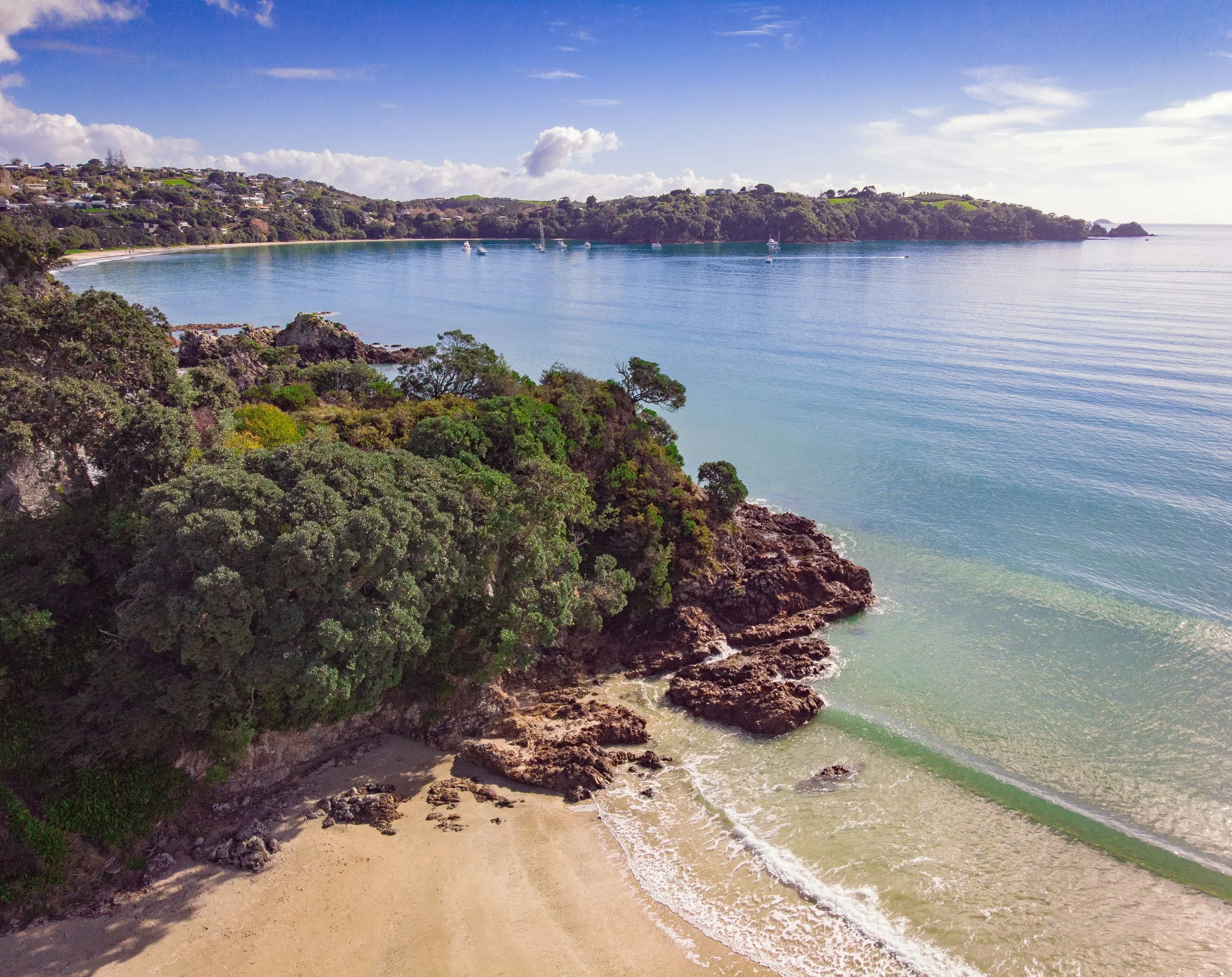 A coastal scene with a sandy beach, rocky cliffs, lush green trees, and calm blue water with a few sailboats and yachts, under a partly cloudy sky.