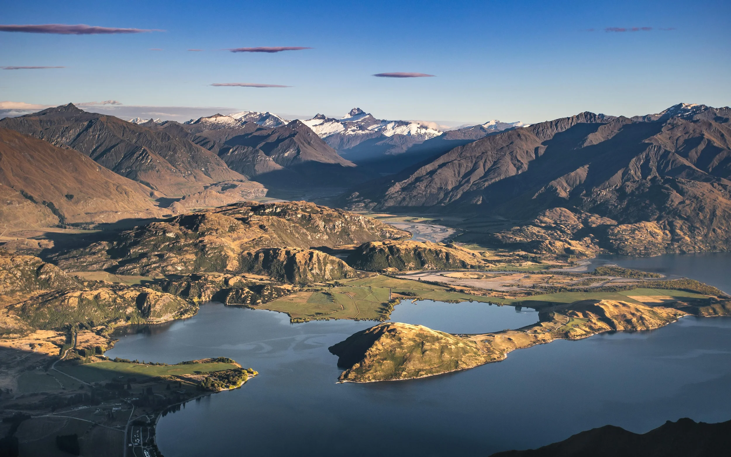 A panoramic view of mountains with snow-capped peaks, a meandering river, and lakes in a valley under a blue sky with a few clouds.