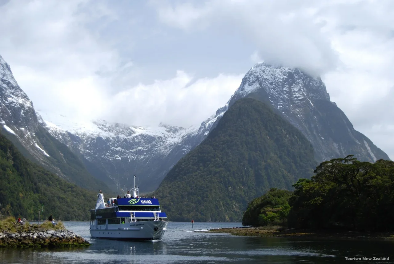 A boat cruising through a fjord with lush green mountains and snow-capped peaks in New Zealand.