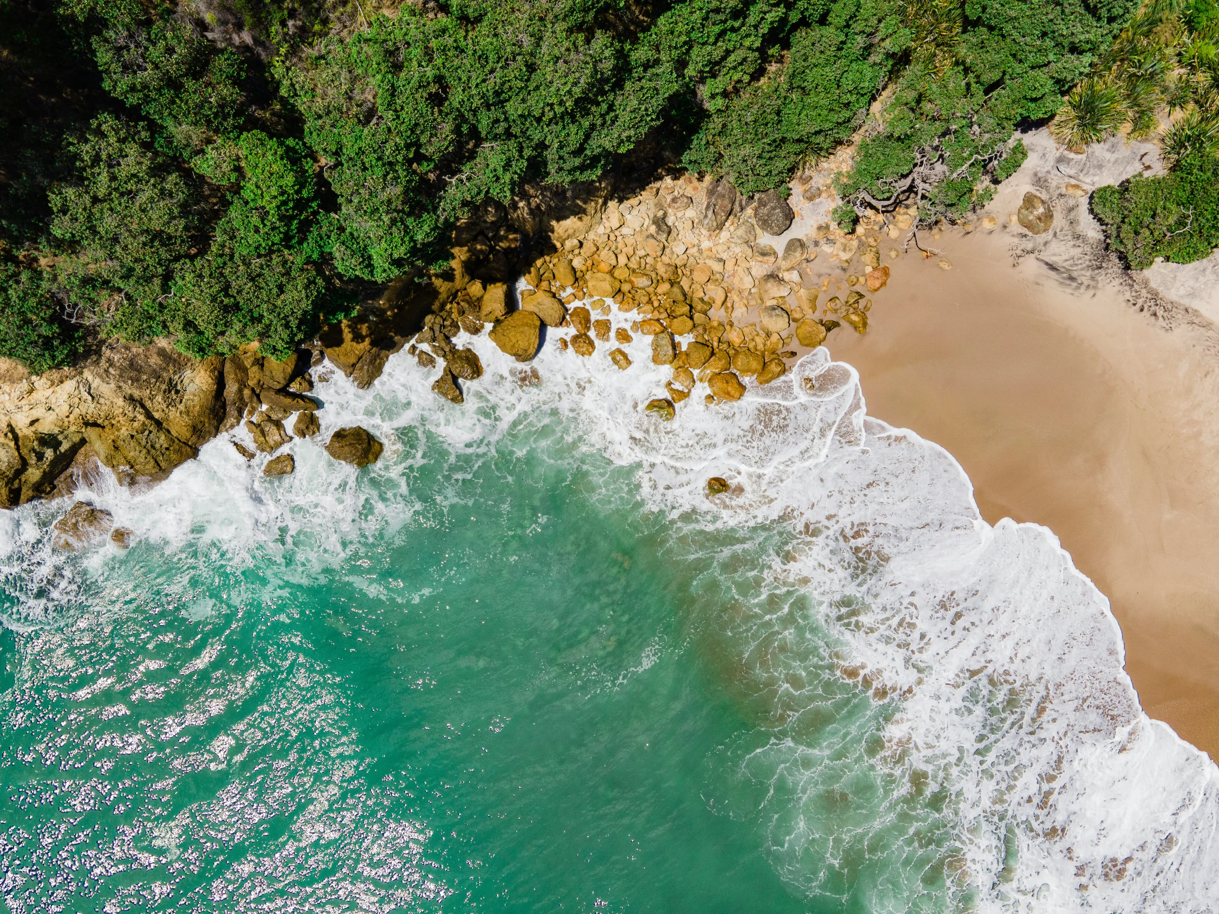 An aerial view of a coastline showing turquoise ocean waters, waves crashing onto sandy beaches, scattered rocks, and dense green forested areas.