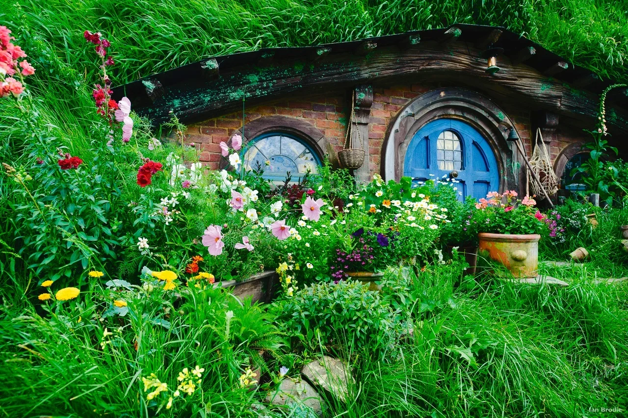 A lush garden scene featuring a Hobbit-style house with a round blue door, surrounded by colorful flowers and green foliage.