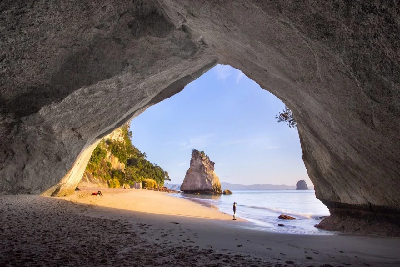 View from inside a beach cave looking out to sandy shore, ocean, and rock formations, with a person standing near the water.