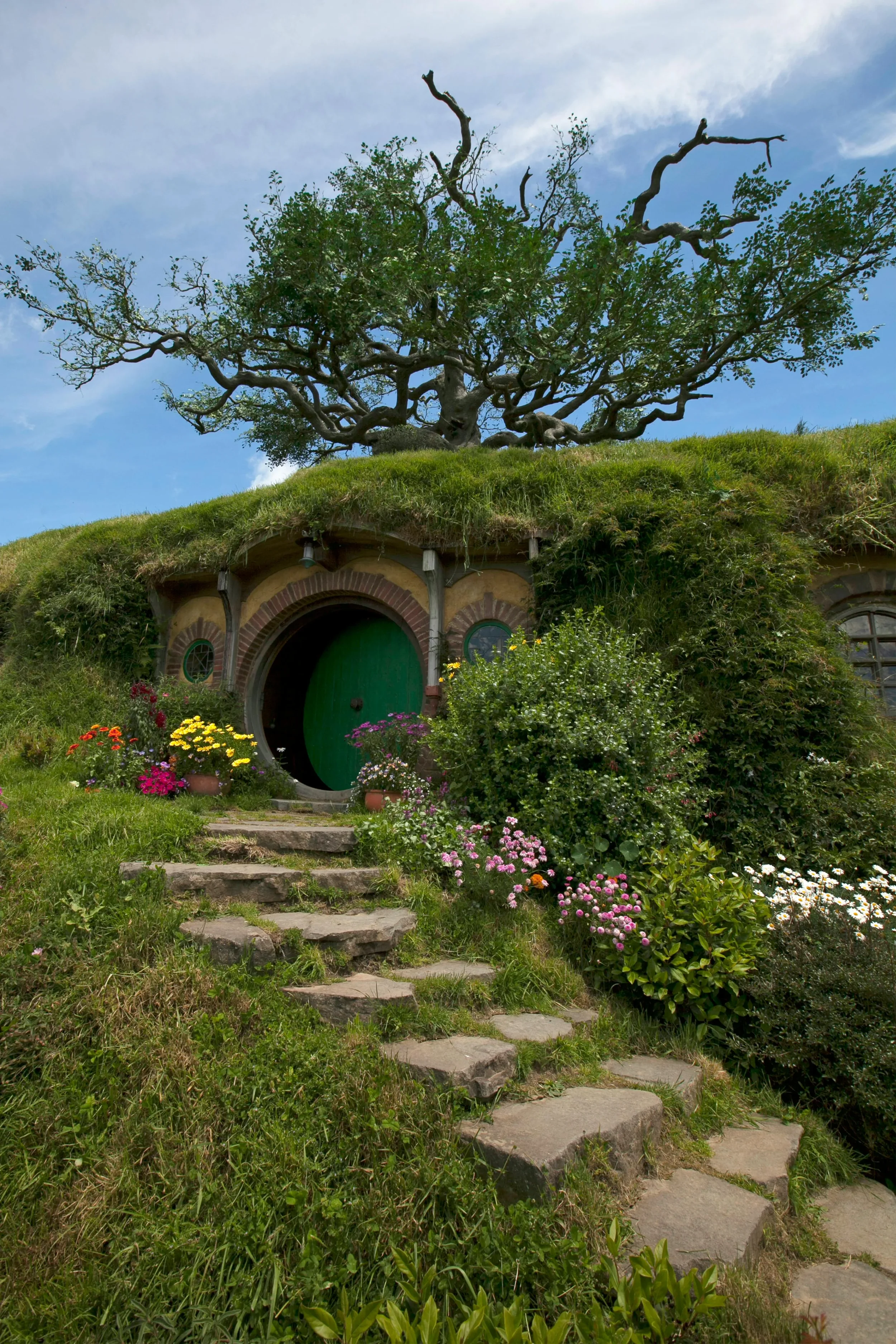 A grassy hill with a large, twisted tree on top and a hobbit hole door at the base. There are flowers along the stone steps leading to the door, and a blue sky with some clouds overhead.