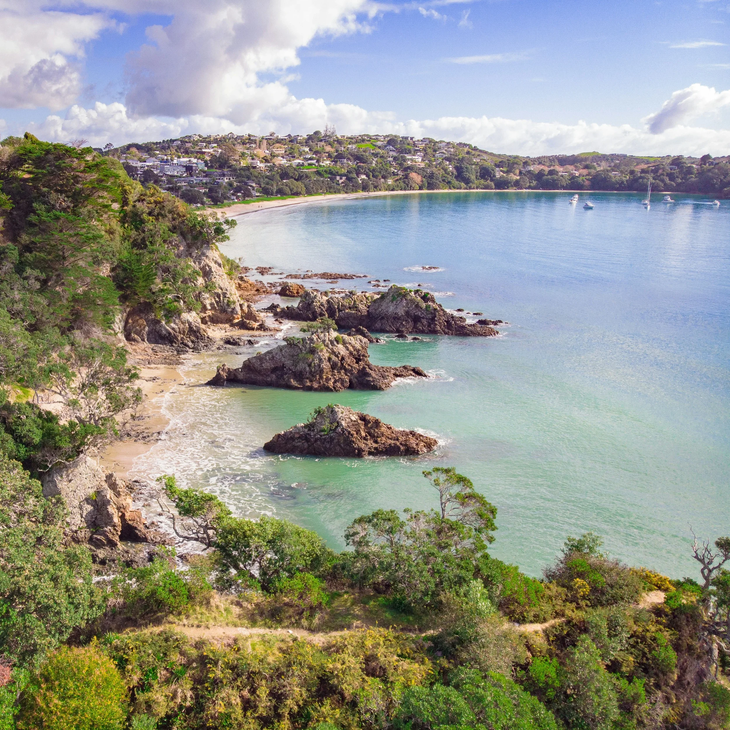 Coastal landscape with a hillside covered in houses, green vegetation, a sandy beach, and boats anchored in the calm water under a partly cloudy sky.