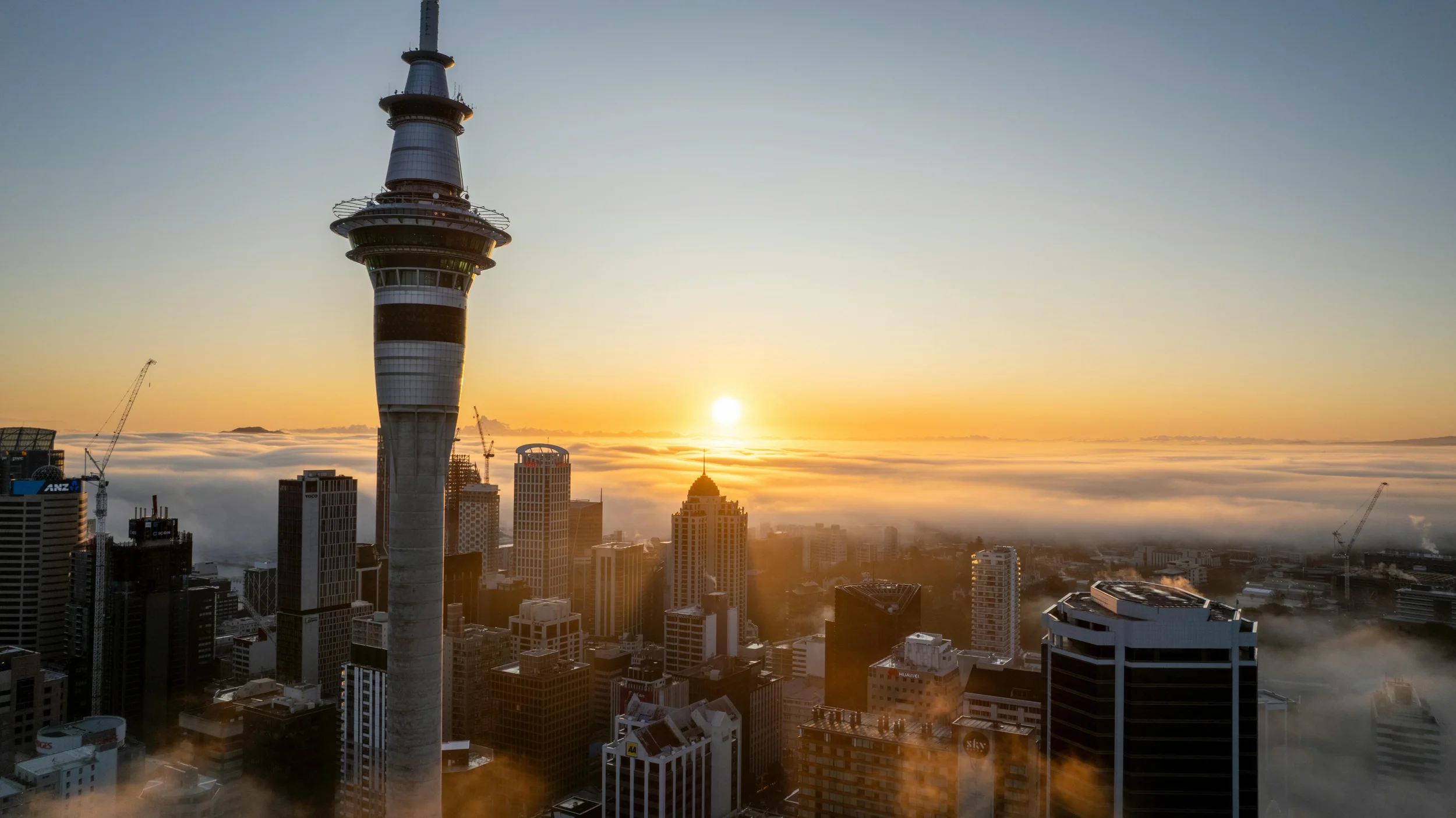 A city skyline at sunrise with tall buildings, cranes, and a tower with viewing platforms, low clouds, and the sun on the horizon.