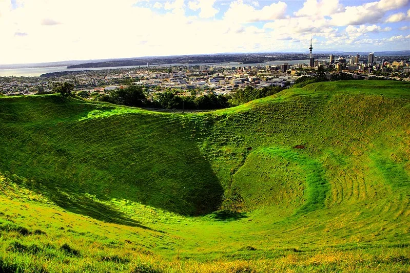 A large, green volcanic crater with a cityscape in the background, including tall buildings and a tower, under a partly cloudy sky.