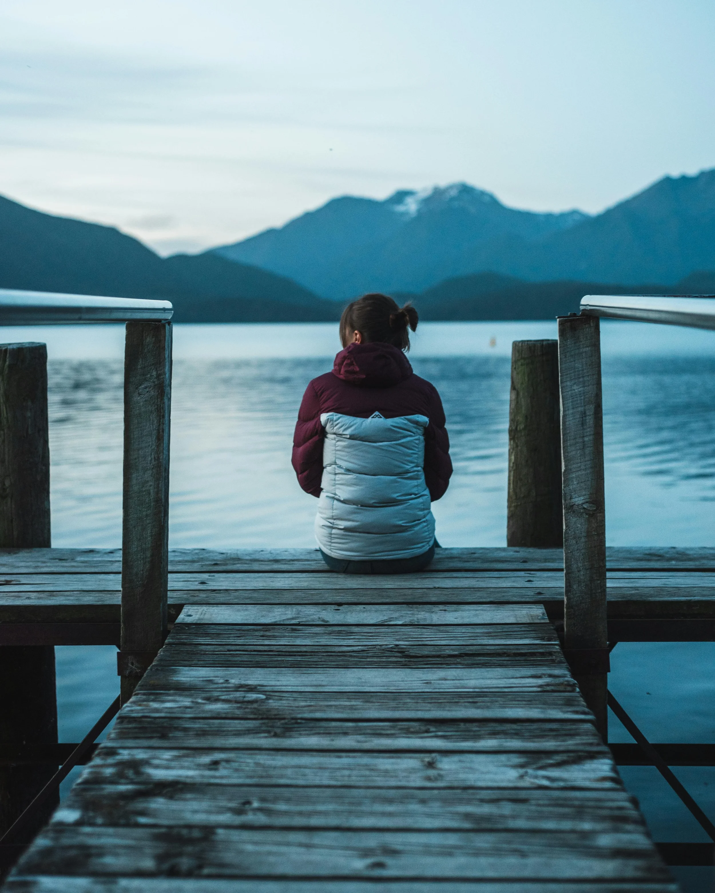 A person with dark hair tied in a bun sitting on a wooden dock overlooking a body of water with mountains in the distance during dusk