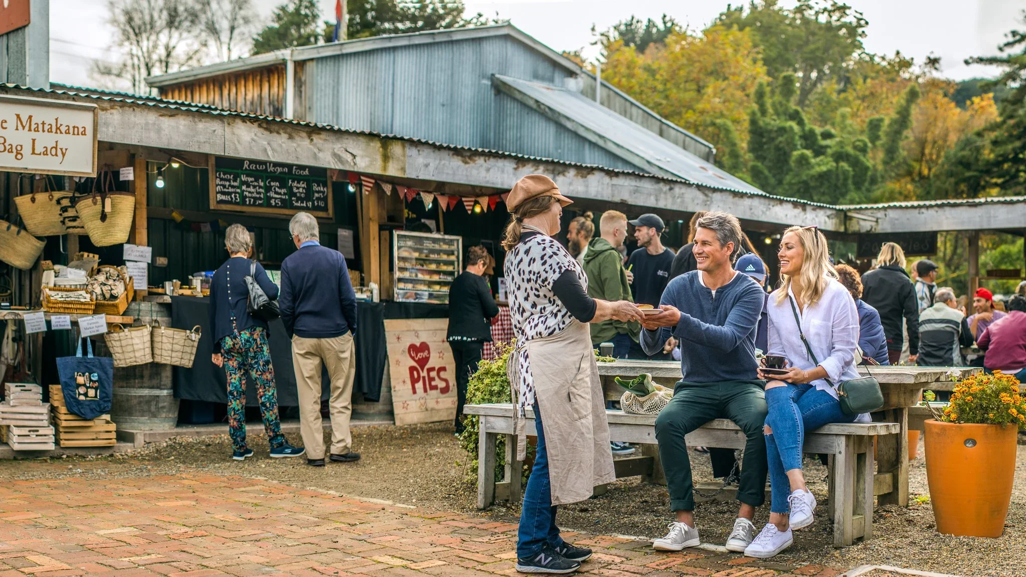 People enjoying a day at an outdoor market with food stalls and vendors, surrounded by trees with fall foliage.