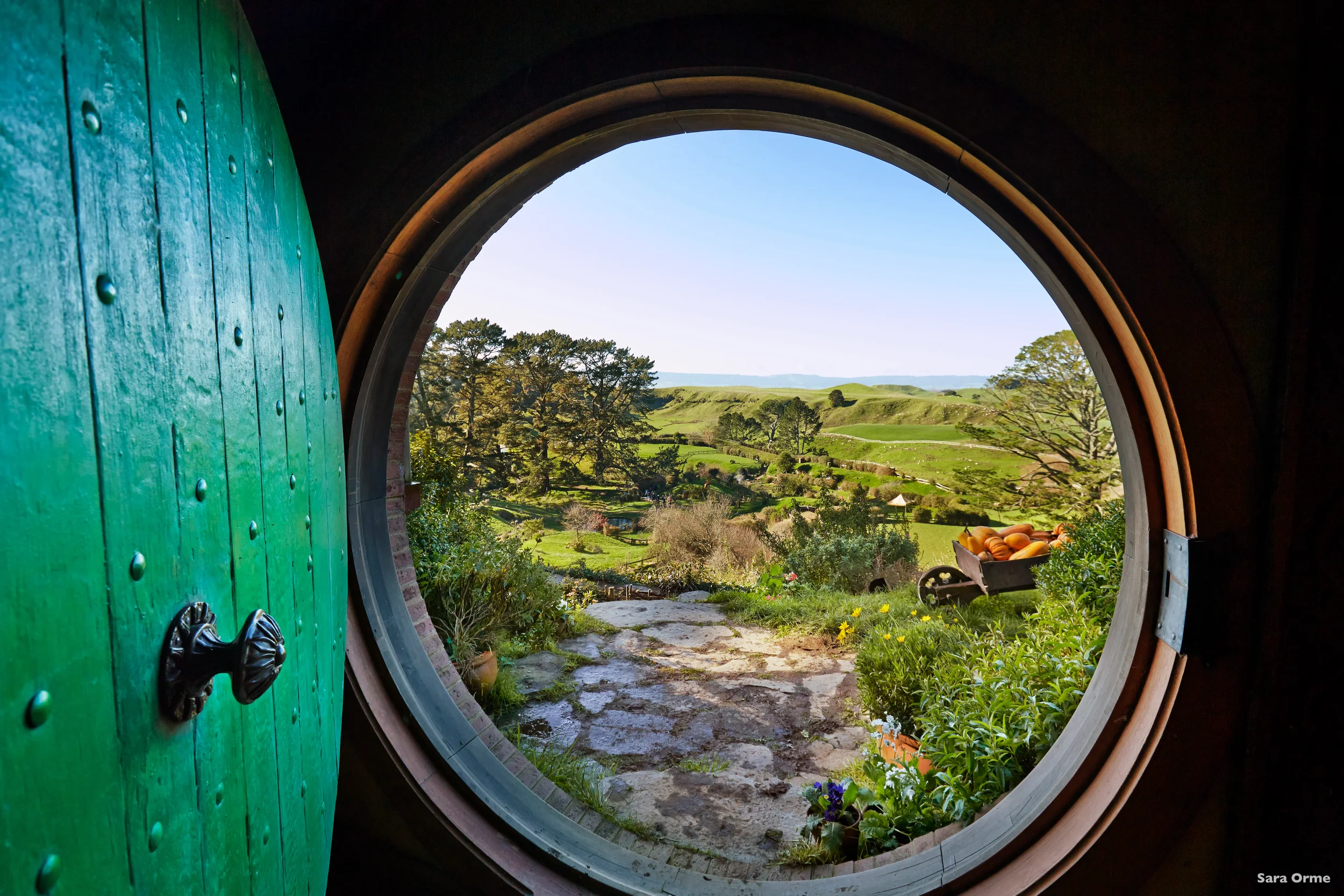 View of a scenic countryside landscape through a Hobbit-style circular door, with lush green hills, trees, and a wheelbarrow filled with pumpkins and flowers outside the door.