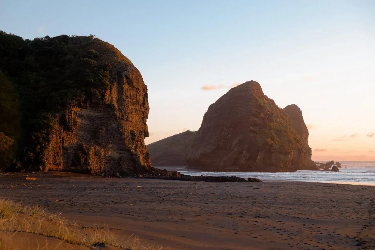 A serene beach scene at sunset with large rocky cliffs and islands in the ocean, sandy shore in the foreground, and a partly cloudy sky.