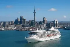 A large cruise ship docked in a harbor with a city skyline and a tall observation tower in the background.