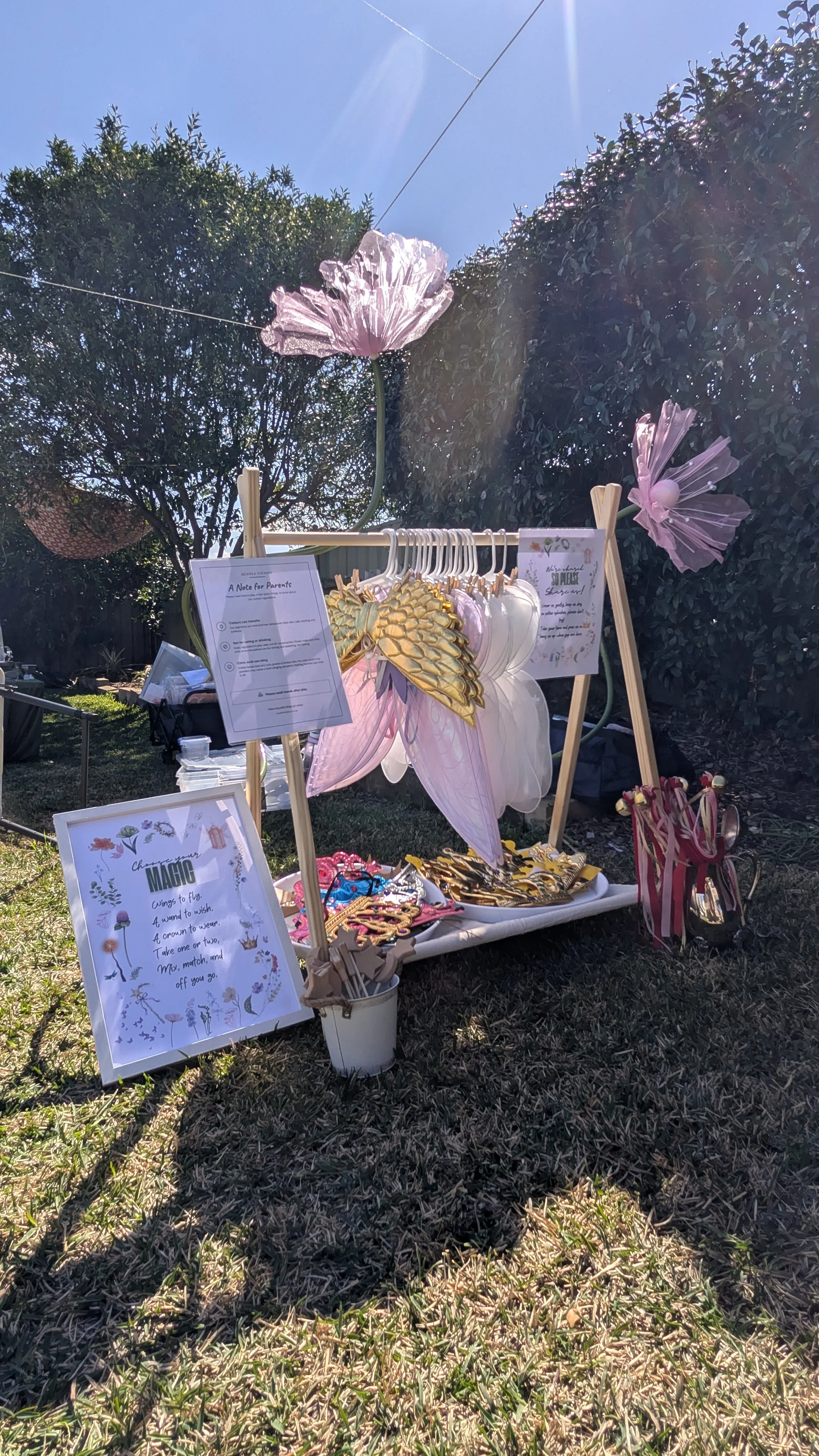 Outdoor display of fairy-themed accessories, including fairy wings, headbands, and decorative signs, set up on a table in a grassy area with trees and a clear sunny sky in the background.