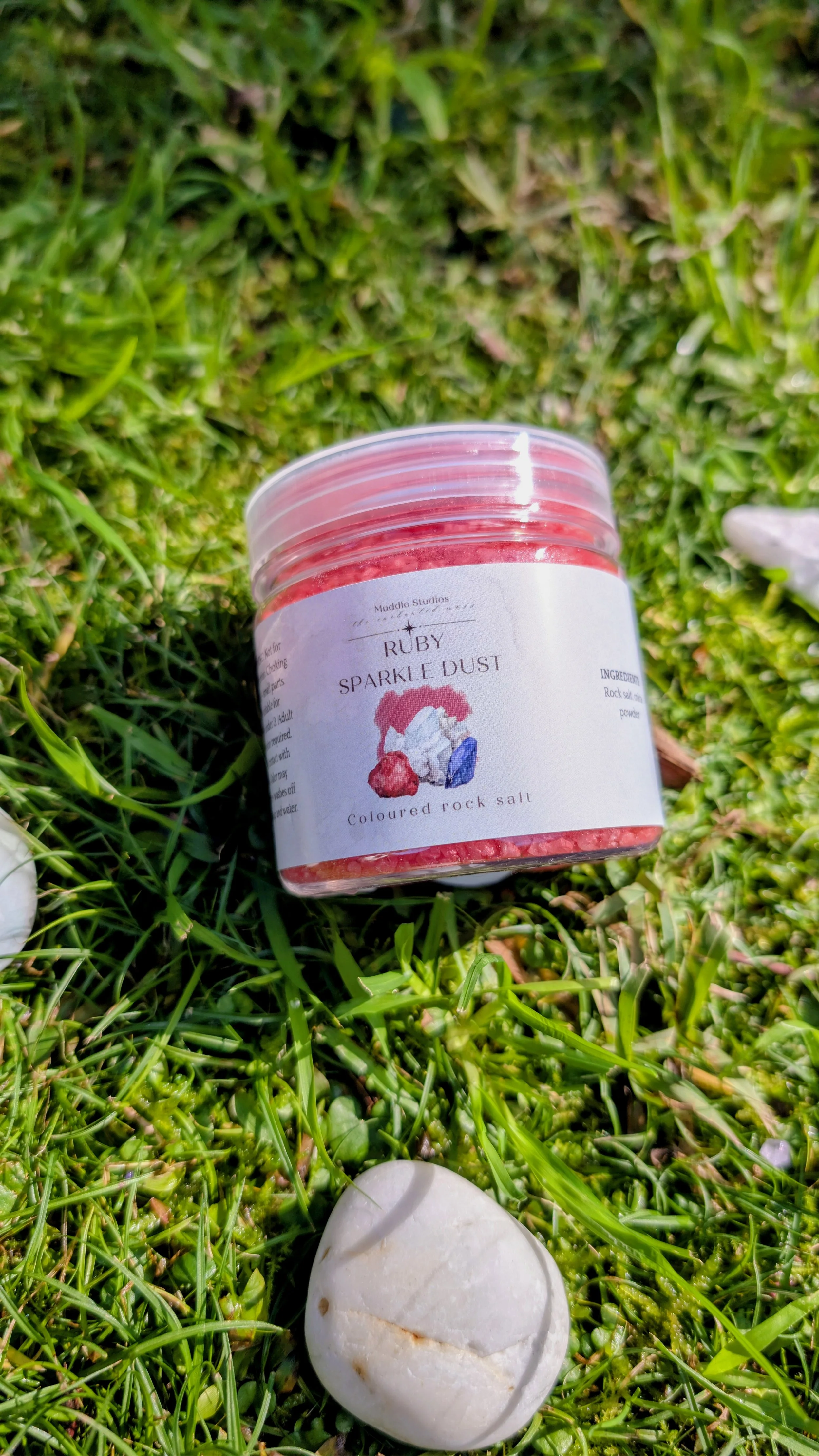 A container labeled 'Ruby Sparkle Dust Coloured rock salt' resting on green grass, with a white rock beside it.
