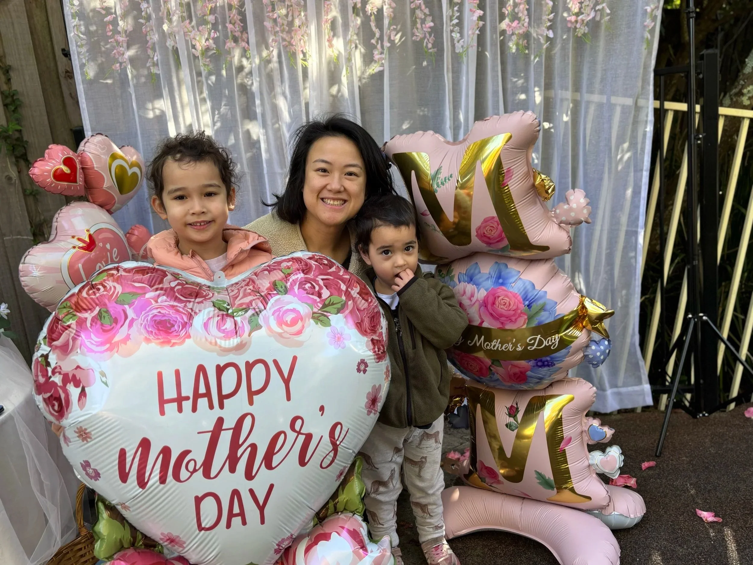 A woman with two young children, all smiling, surrounded by pink and floral balloons, including a large heart-shaped balloon that reads 'Happy Mother's Day' and other balloons with floral designs, in a garden setting with a sheer curtain backdrop.
