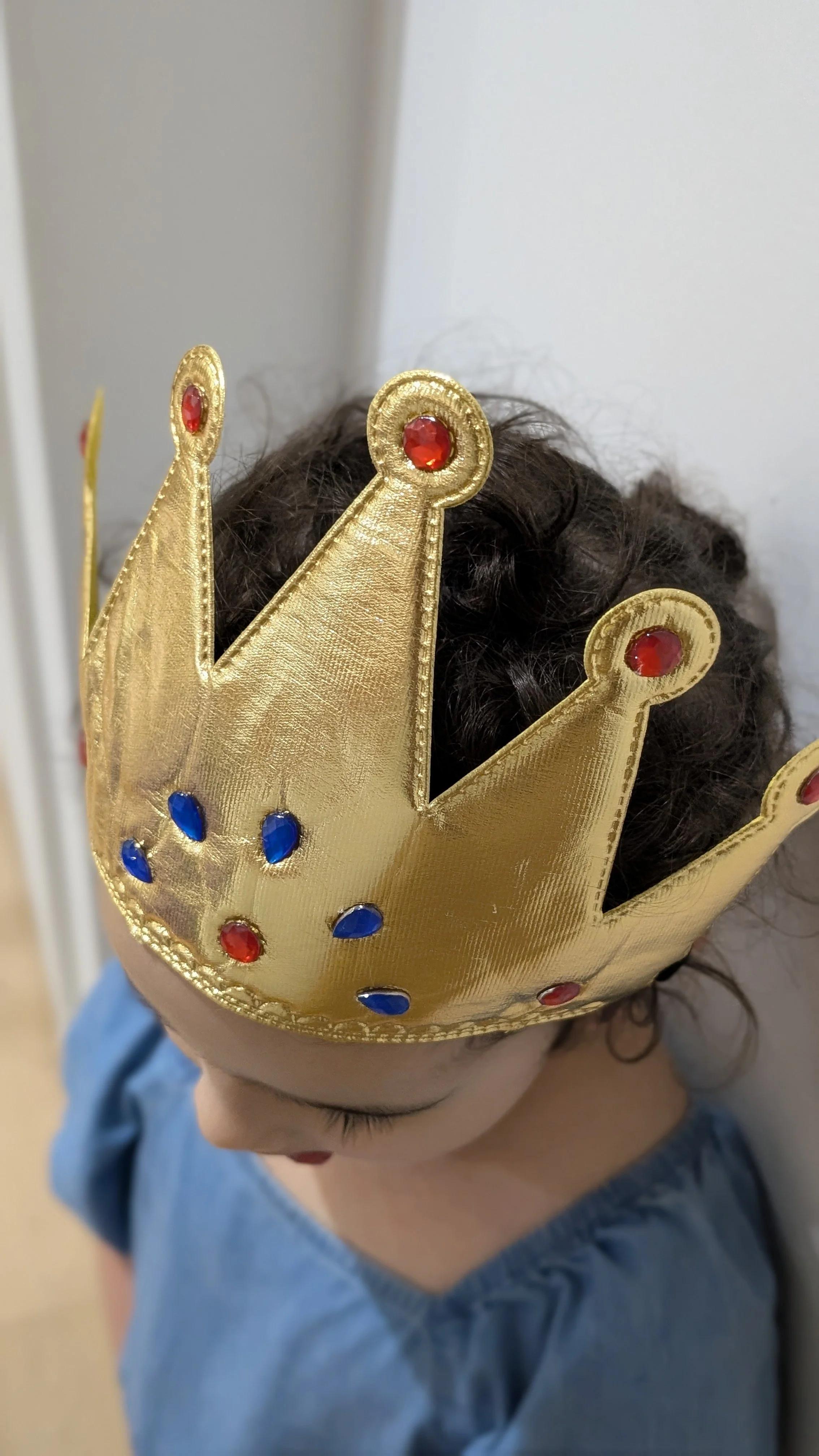 Child wearing a gold crown decorated with red and blue gemstones, looking down with curly hair.