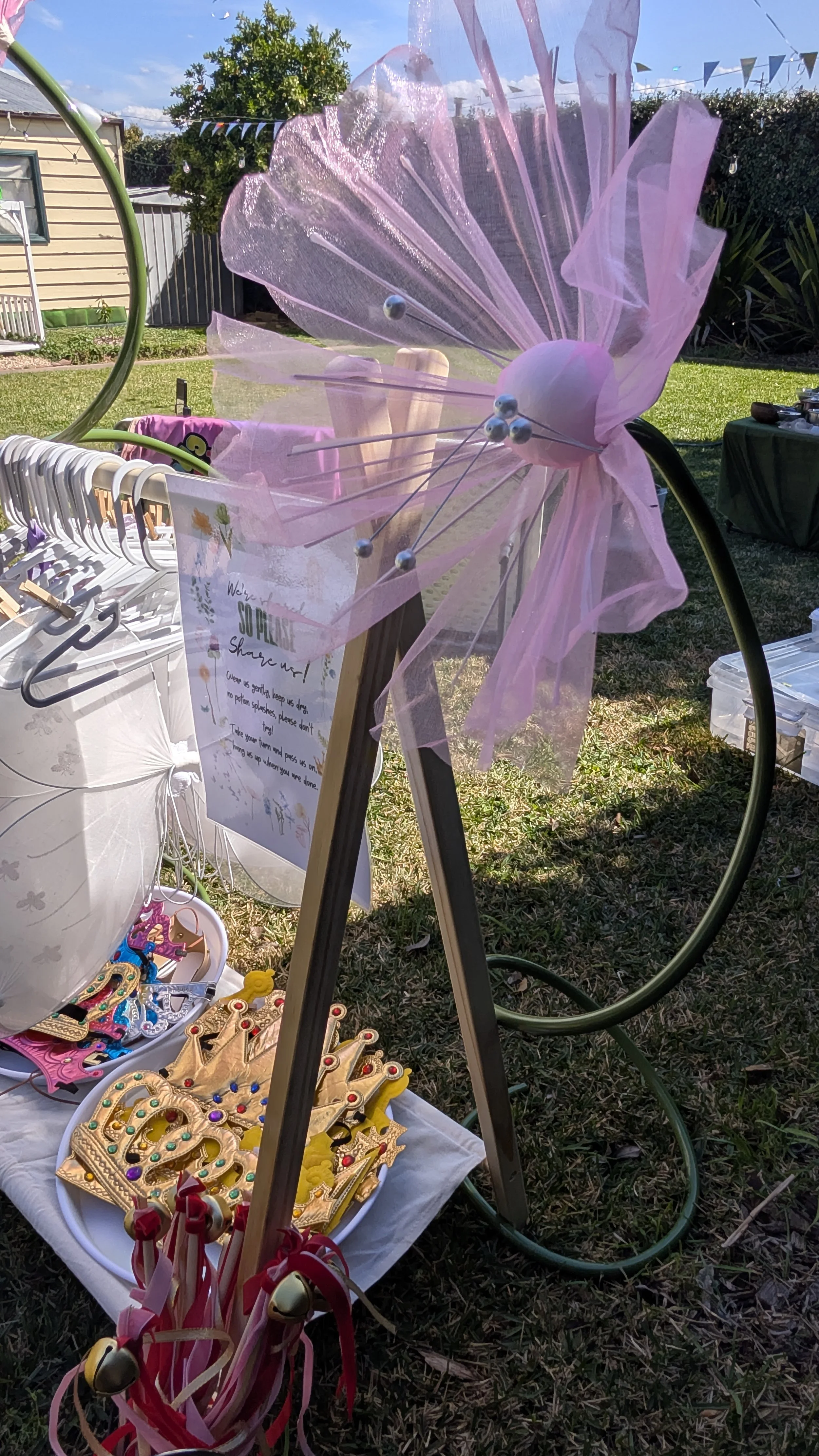 A display table at an outdoor market with gold-colored royal crown jewelry, pink and red ribbon wands, and several decorative items, with a pink and purple fairy wing decoration attached to a metal stand and a sign