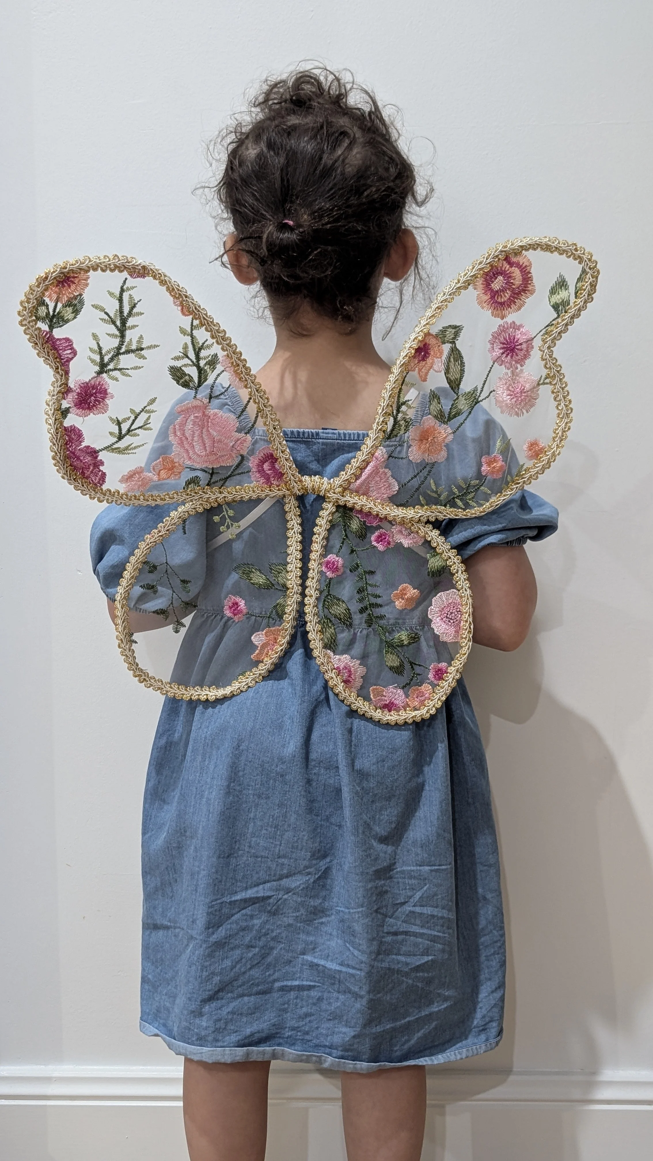 Child dressed in a denim dress with floral embroidered fairy wings standing with their back to the camera against a plain wall.