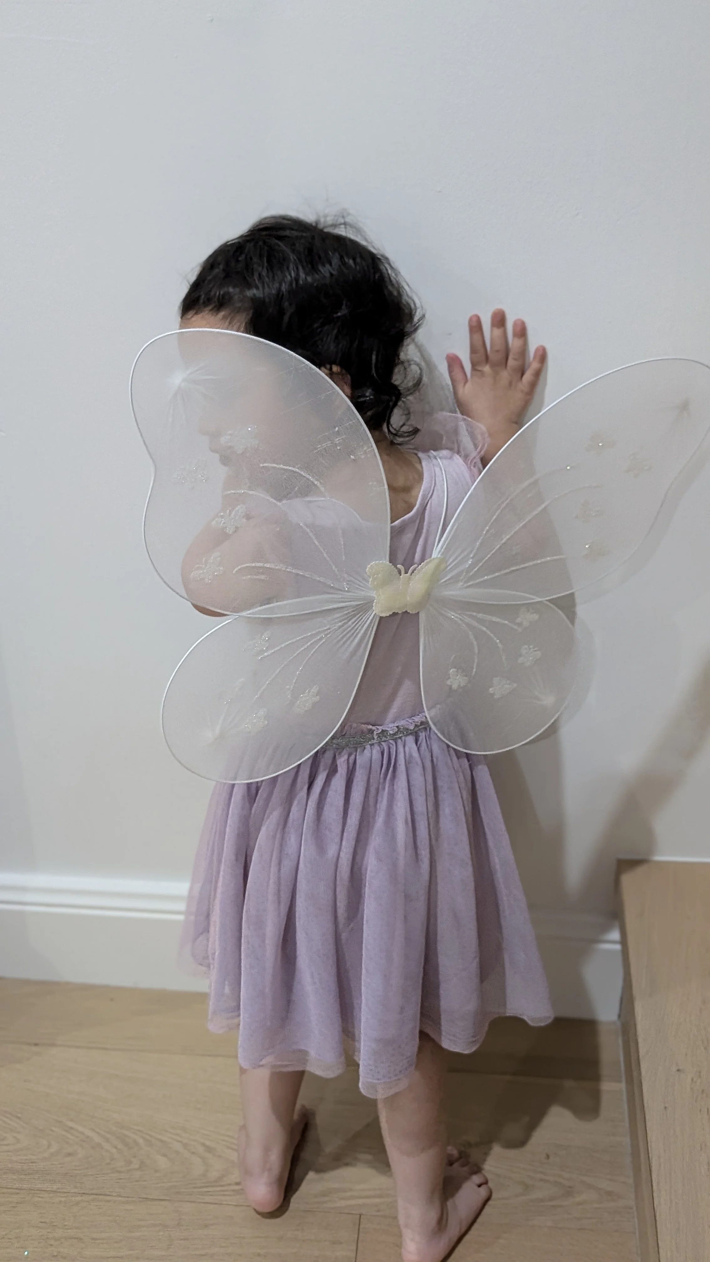 Child dressed in a purple fairy costume with transparent butterfly wings, standing barefoot on a wooden floor against a plain white wall.