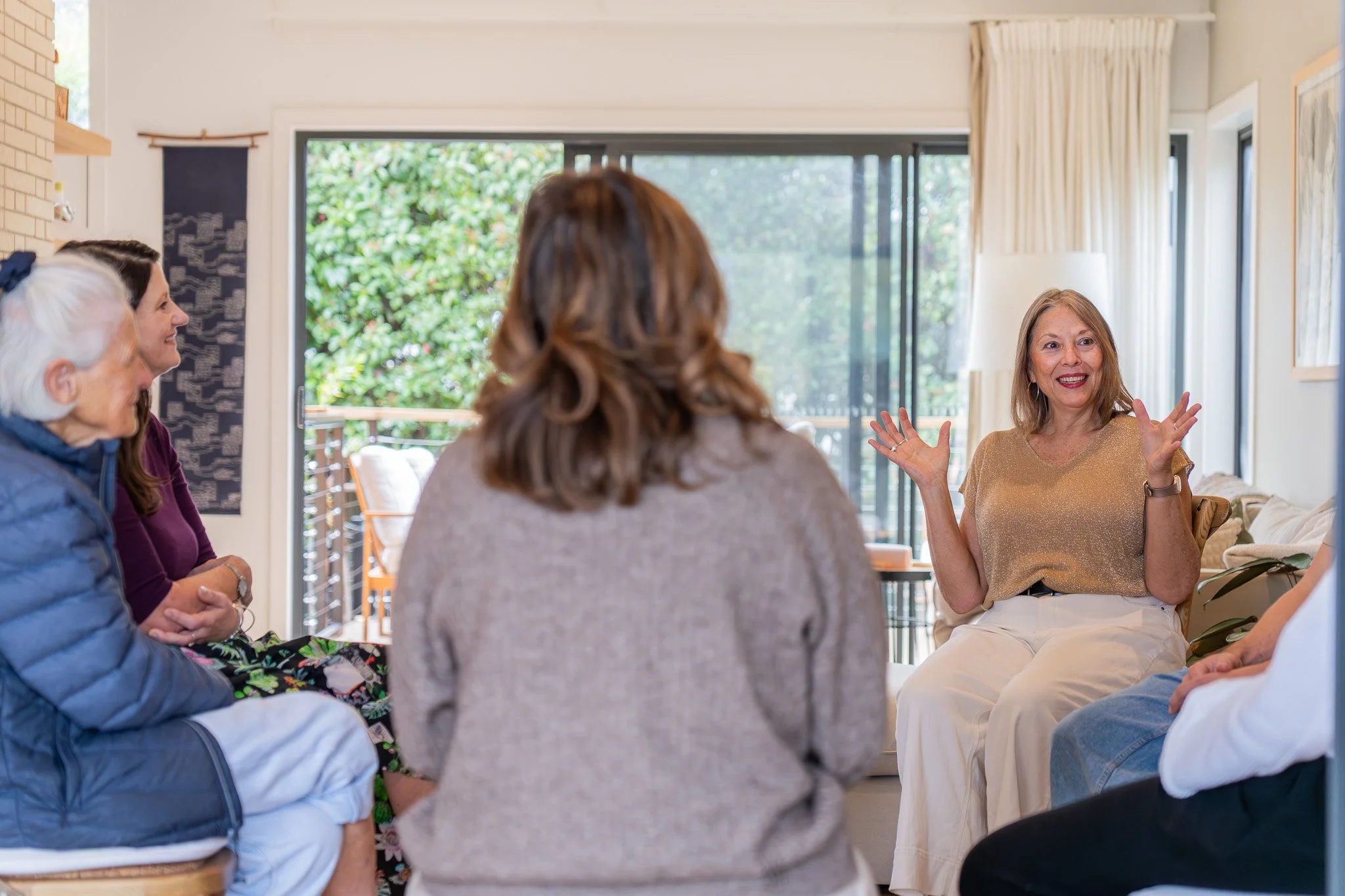 A group of women sitting in a circle and engaging in conversation in a well-lit living room with large windows and greenery outside. A woman on the right is gesturing with her hands while speaking.